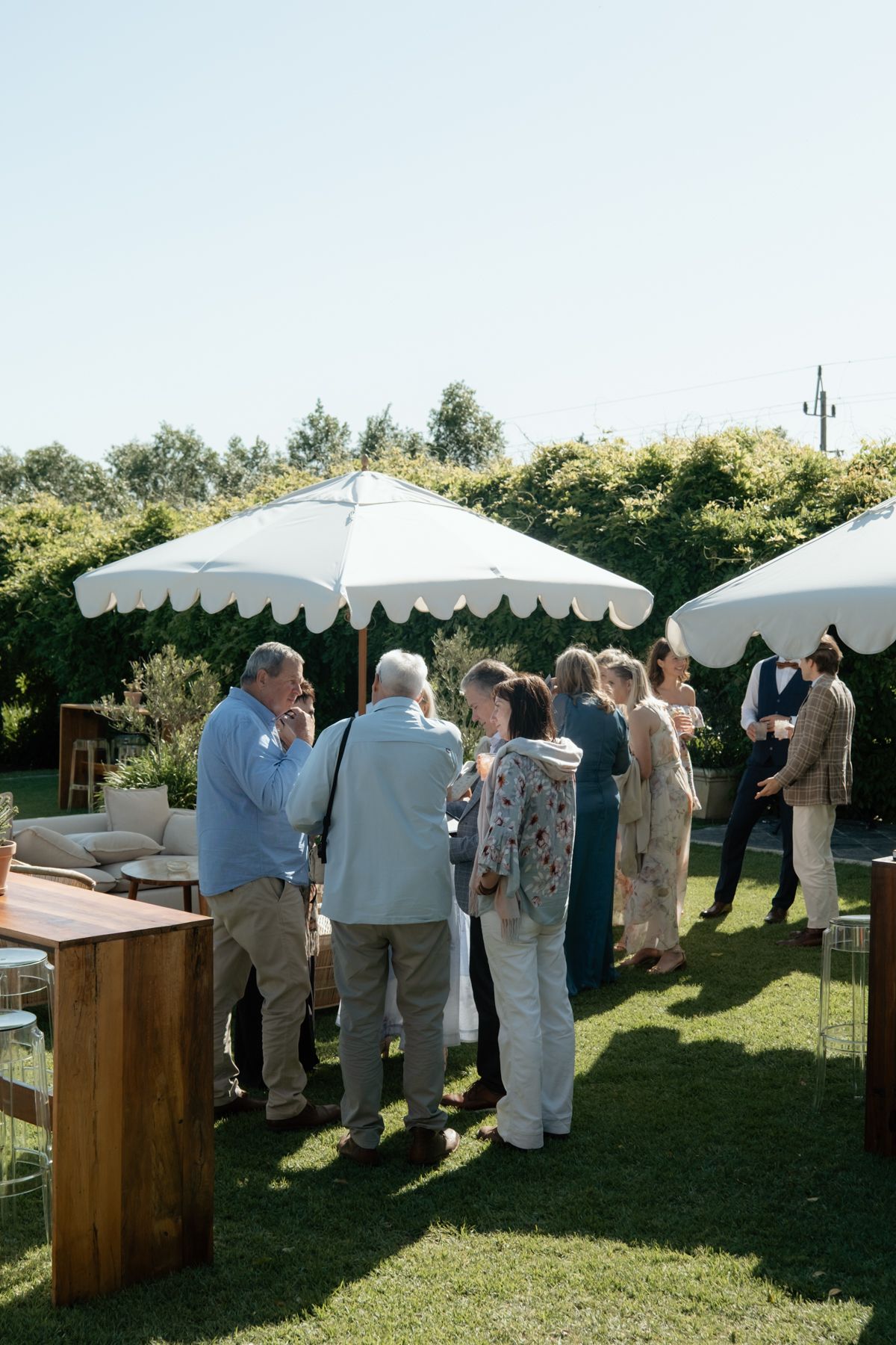Wedding guests mingle under white market umbrellas on sunny outdoor garden reception lawn.