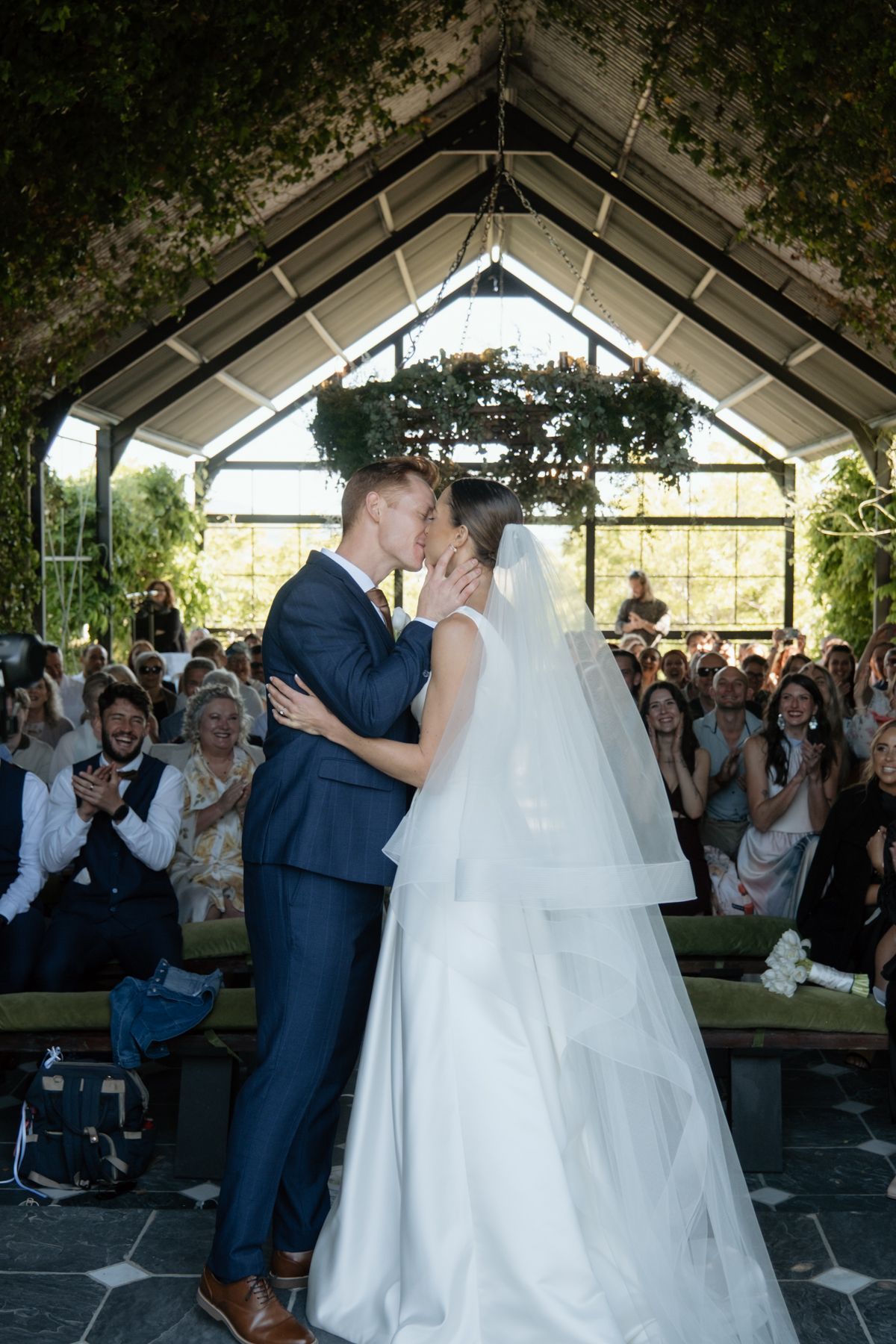 Bride and groom sharing their first kiss under a glass chapel arch before cheering guests.