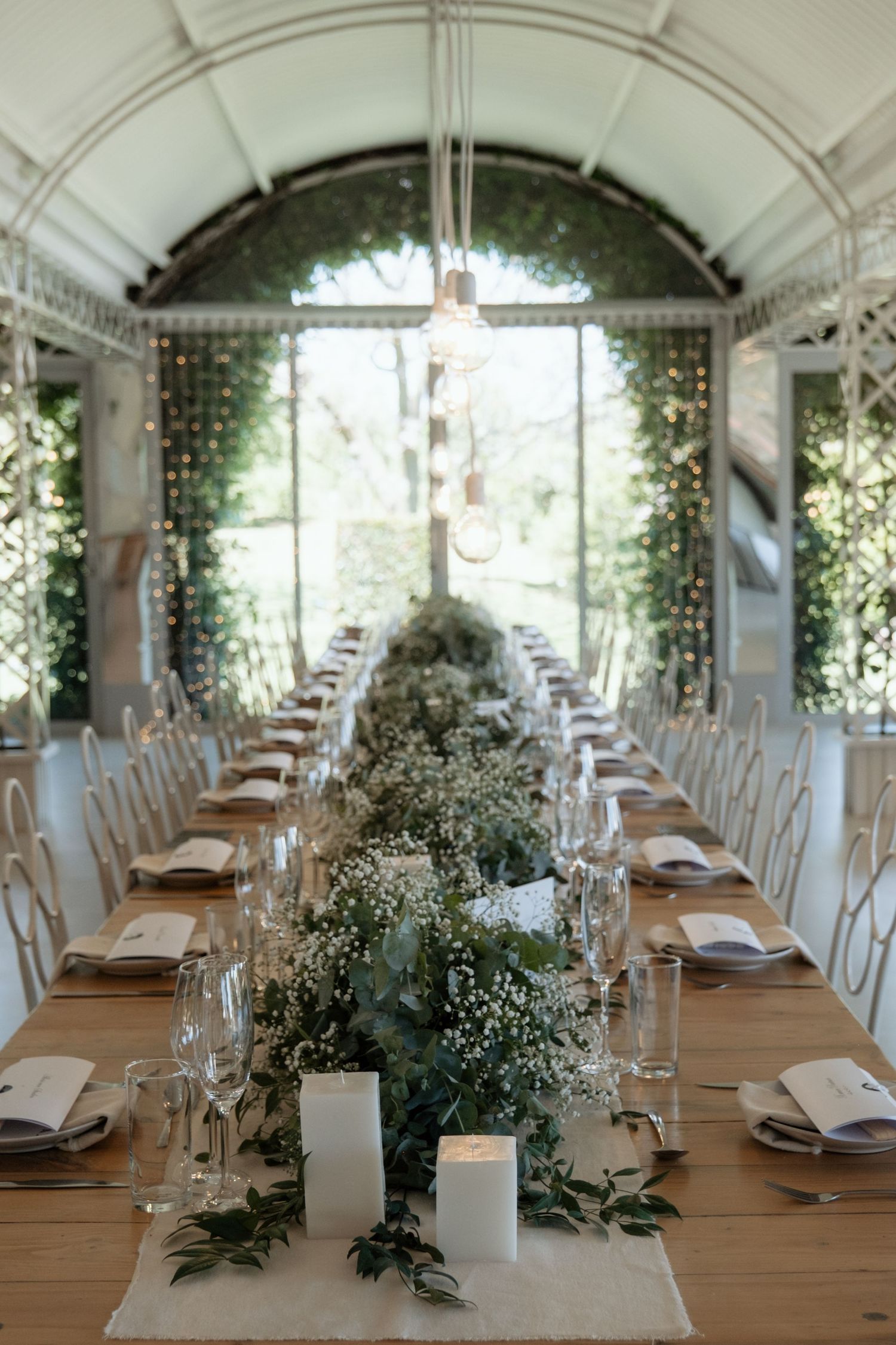 Elegant greenhouse wedding reception with long wooden table adorned with white floral greenery centerpieces.