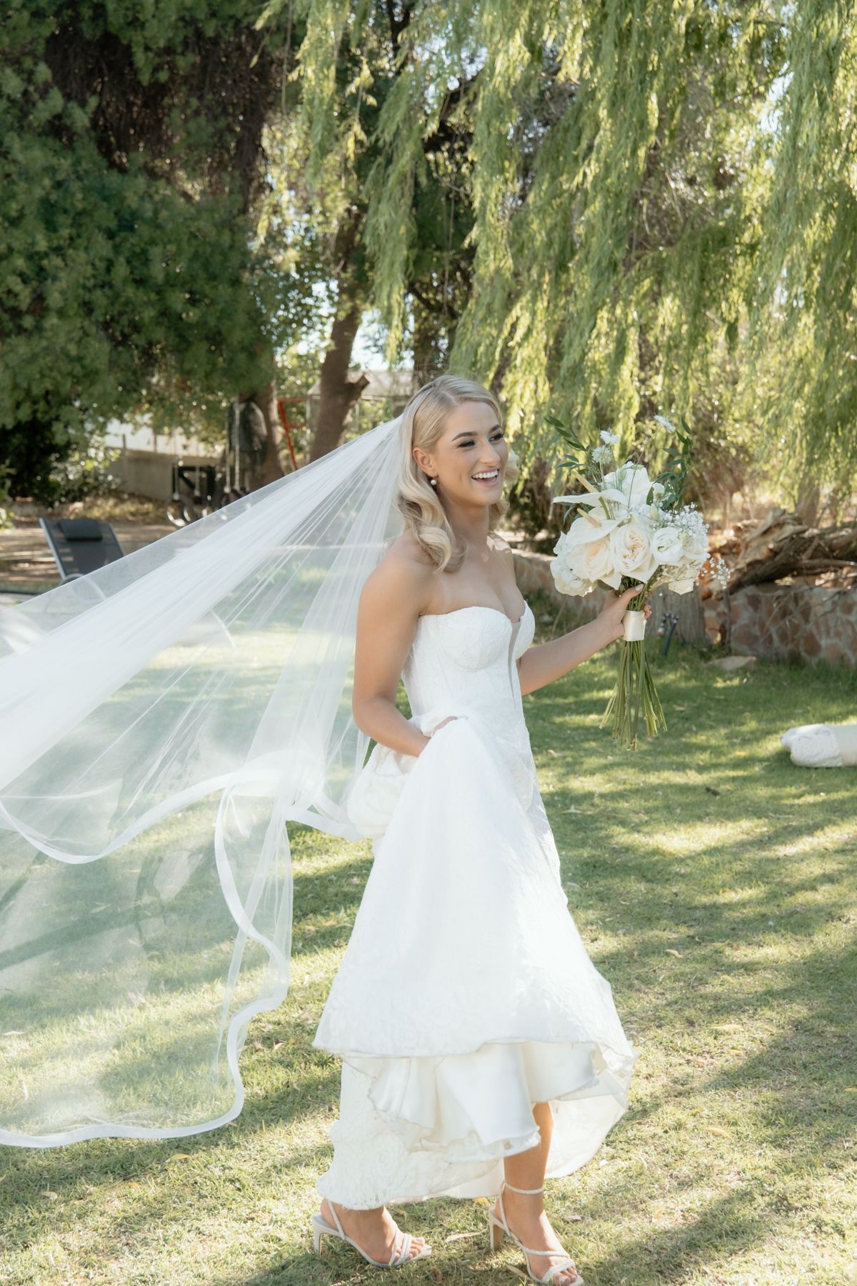 Radiant bride in strapless white gown with flowing veil holds white bouquet under a weeping willow tree.