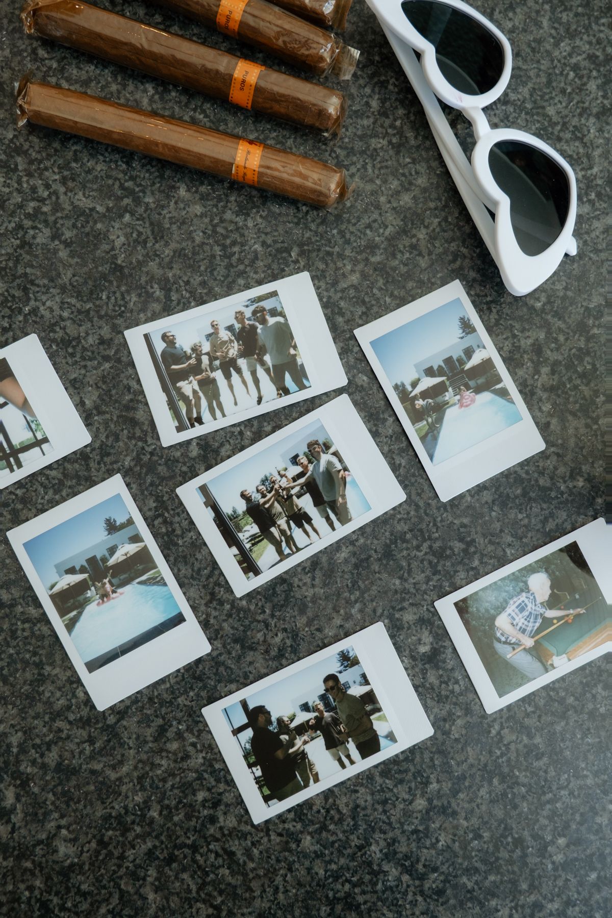 Polaroid photos, cigars and white sunglasses arranged on a dark marble surface at a wedding celebration.