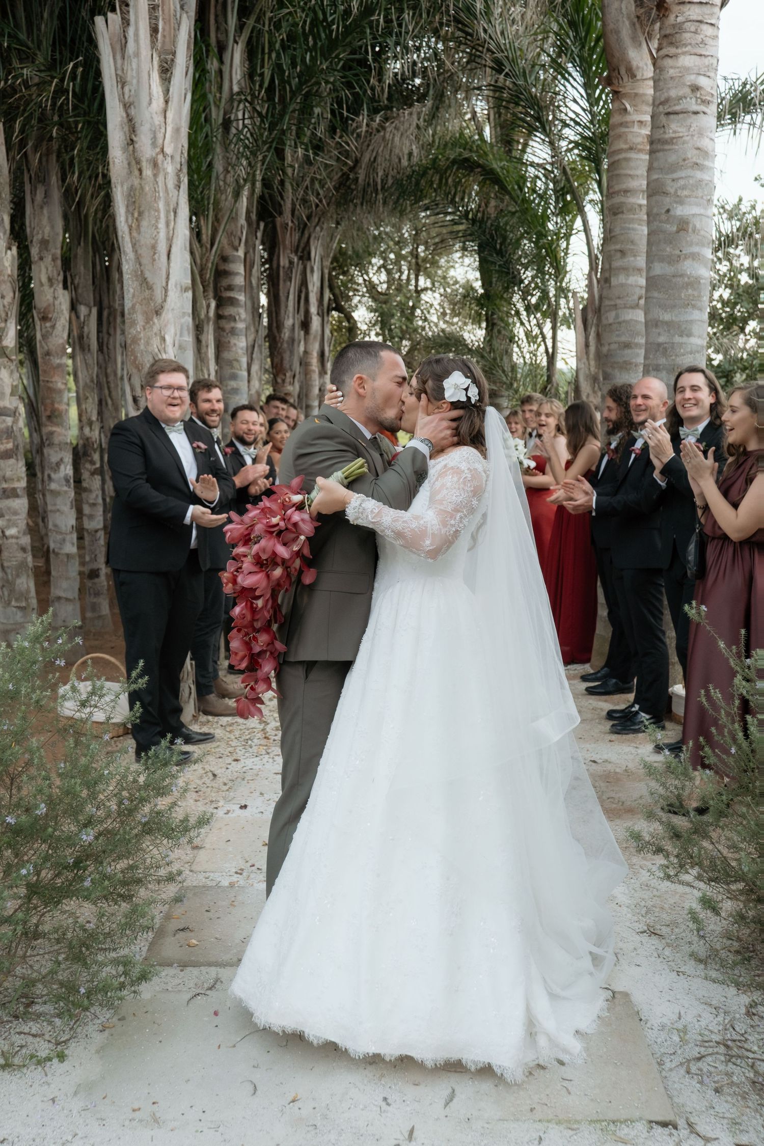 Bride and groom kiss during outdoor wedding recessional lined with guests under palm trees.