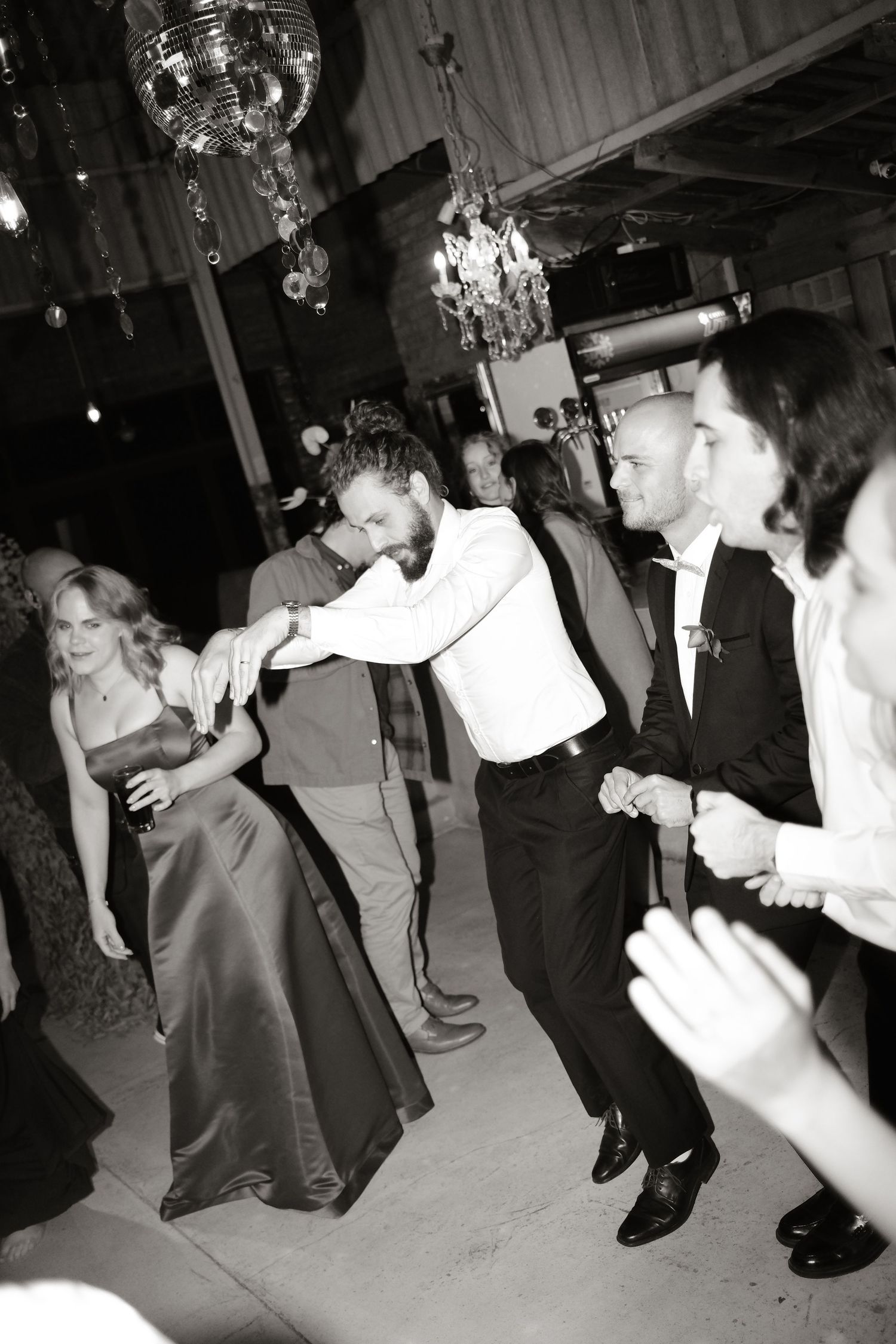 Black and white photo of wedding guests dancing energetically on reception dance floor under chandelier lights.