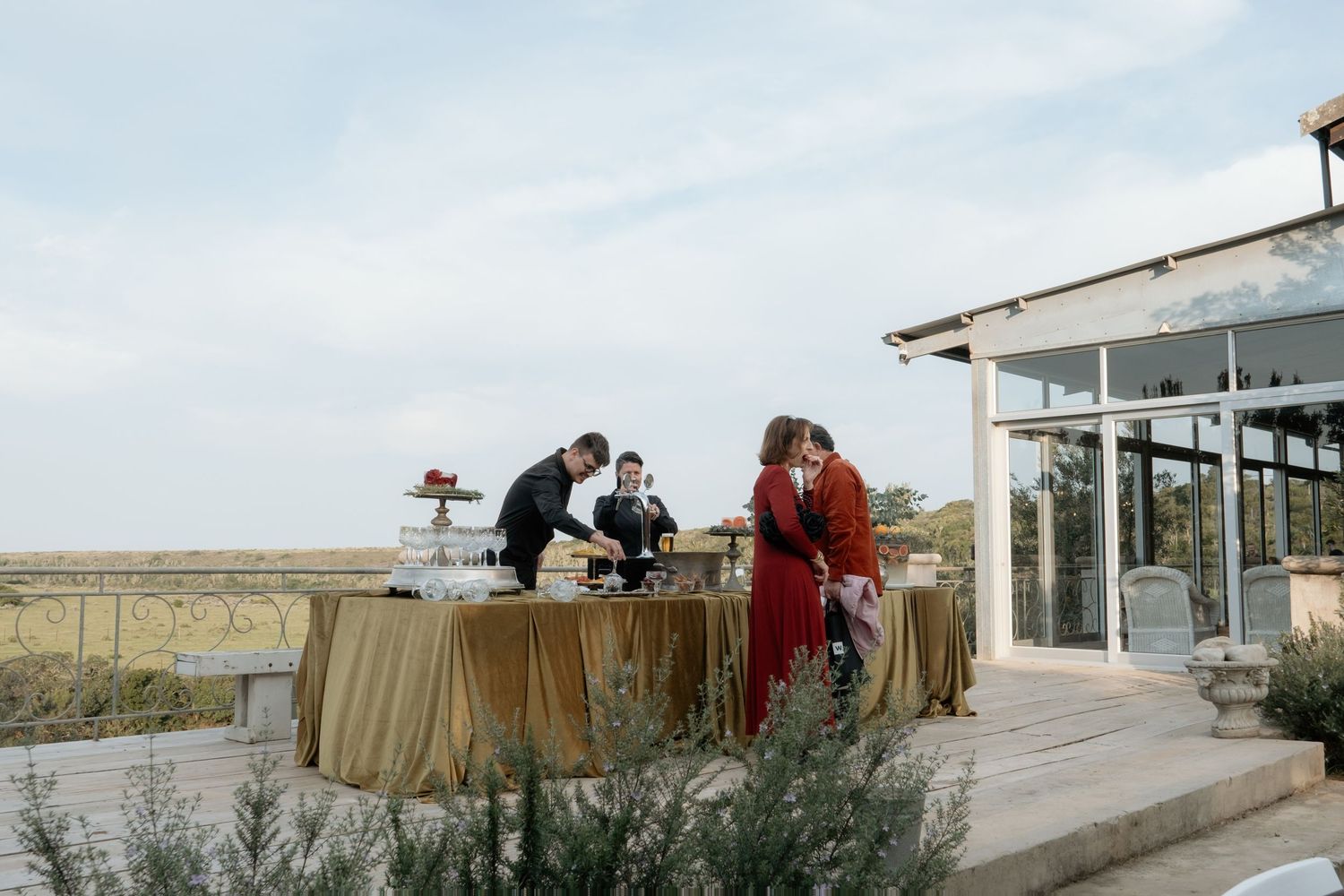 Outdoor wedding cocktail hour with guests at a draped table beside a glass venue building.