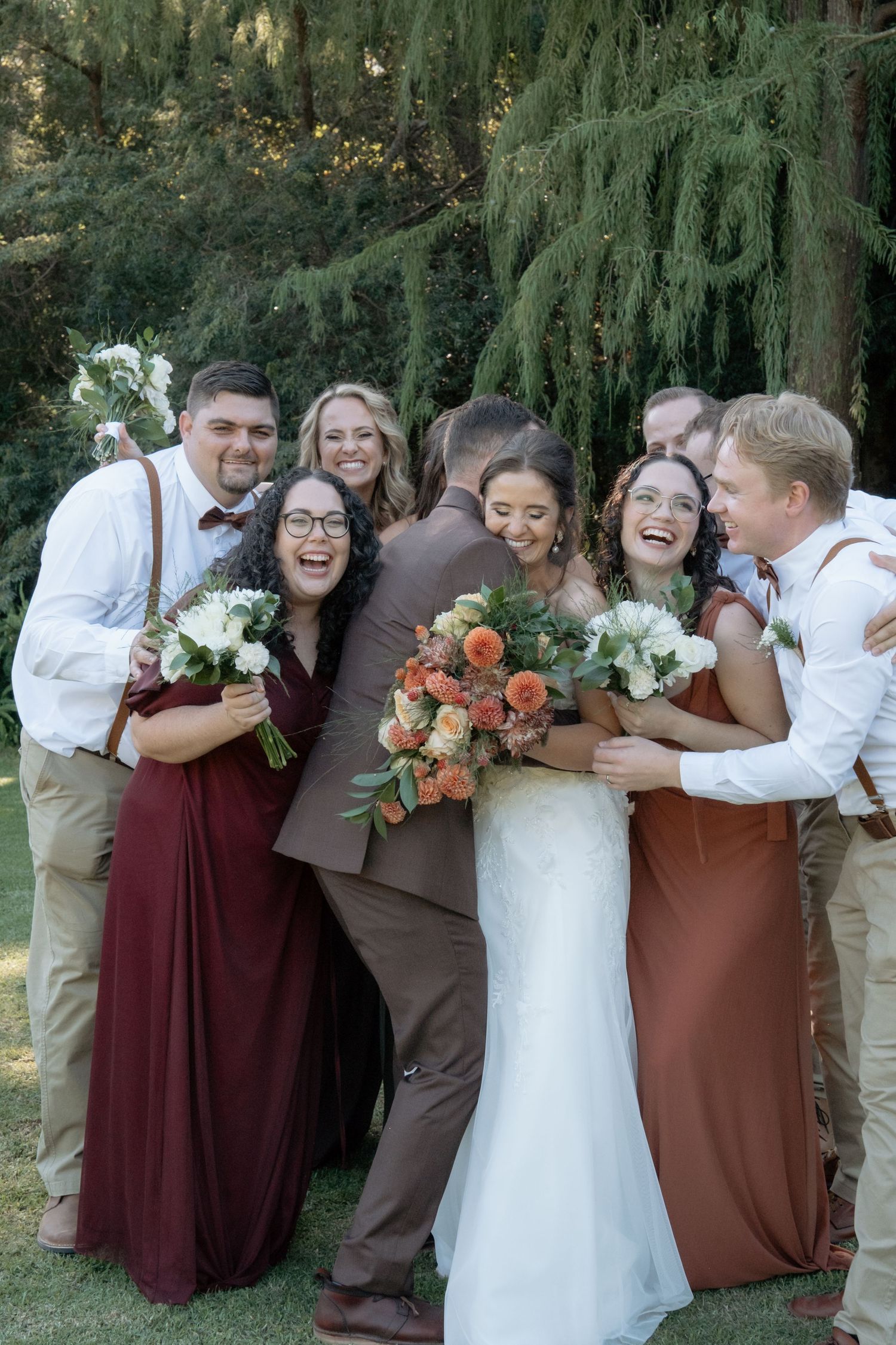 Happy wedding party group hugs bride and groom under tall evergreen trees.