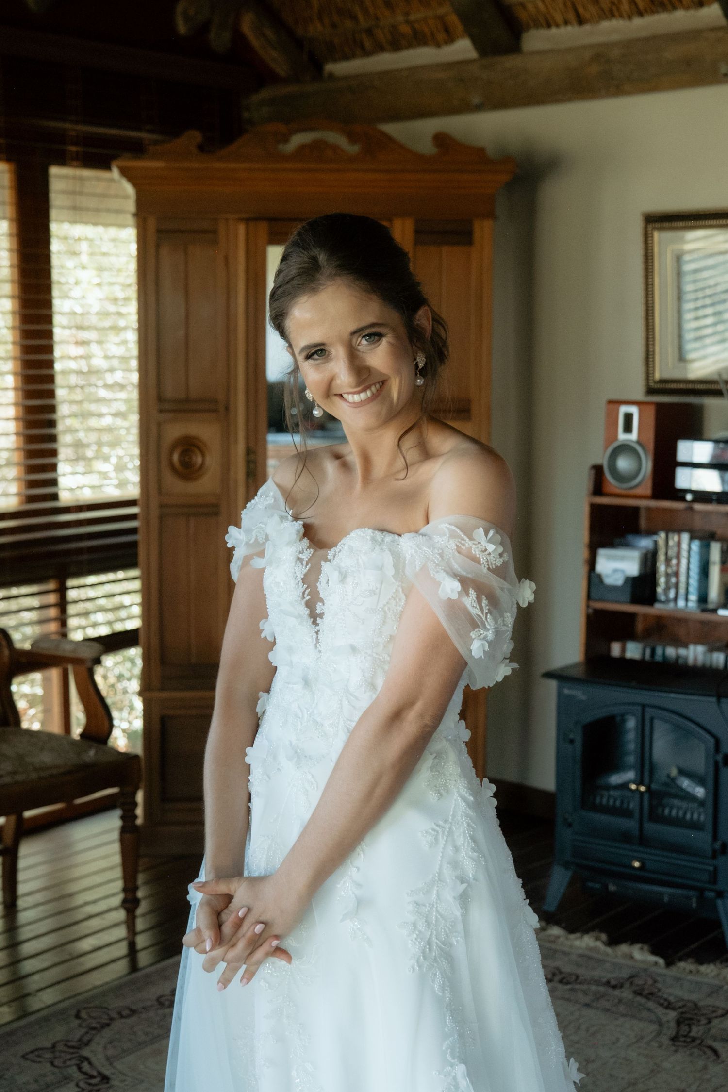 Smiling bride in an off-shoulder floral lace wedding gown posing in a rustic lodge room.