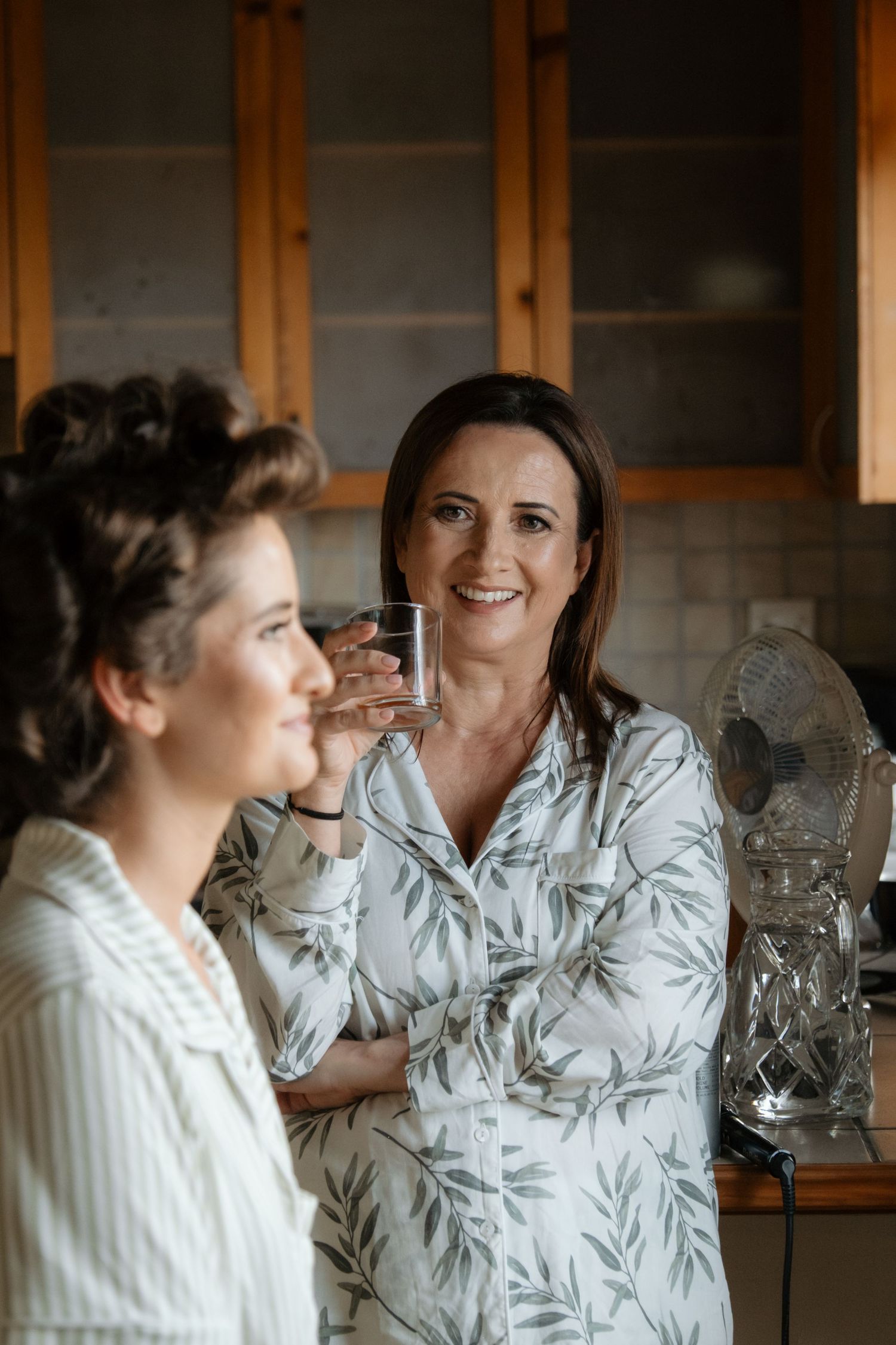 Two women in pajamas chat in kitchen during bridal morning preparations, one holding water glass.
