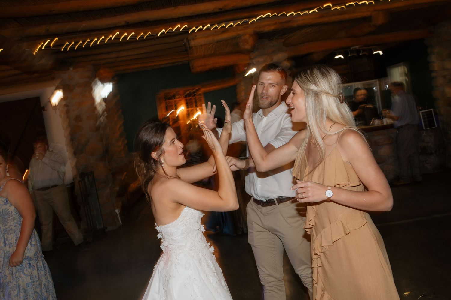 Bride in white dress takes photo of guests dancing at rustic wedding reception with warm string lights.