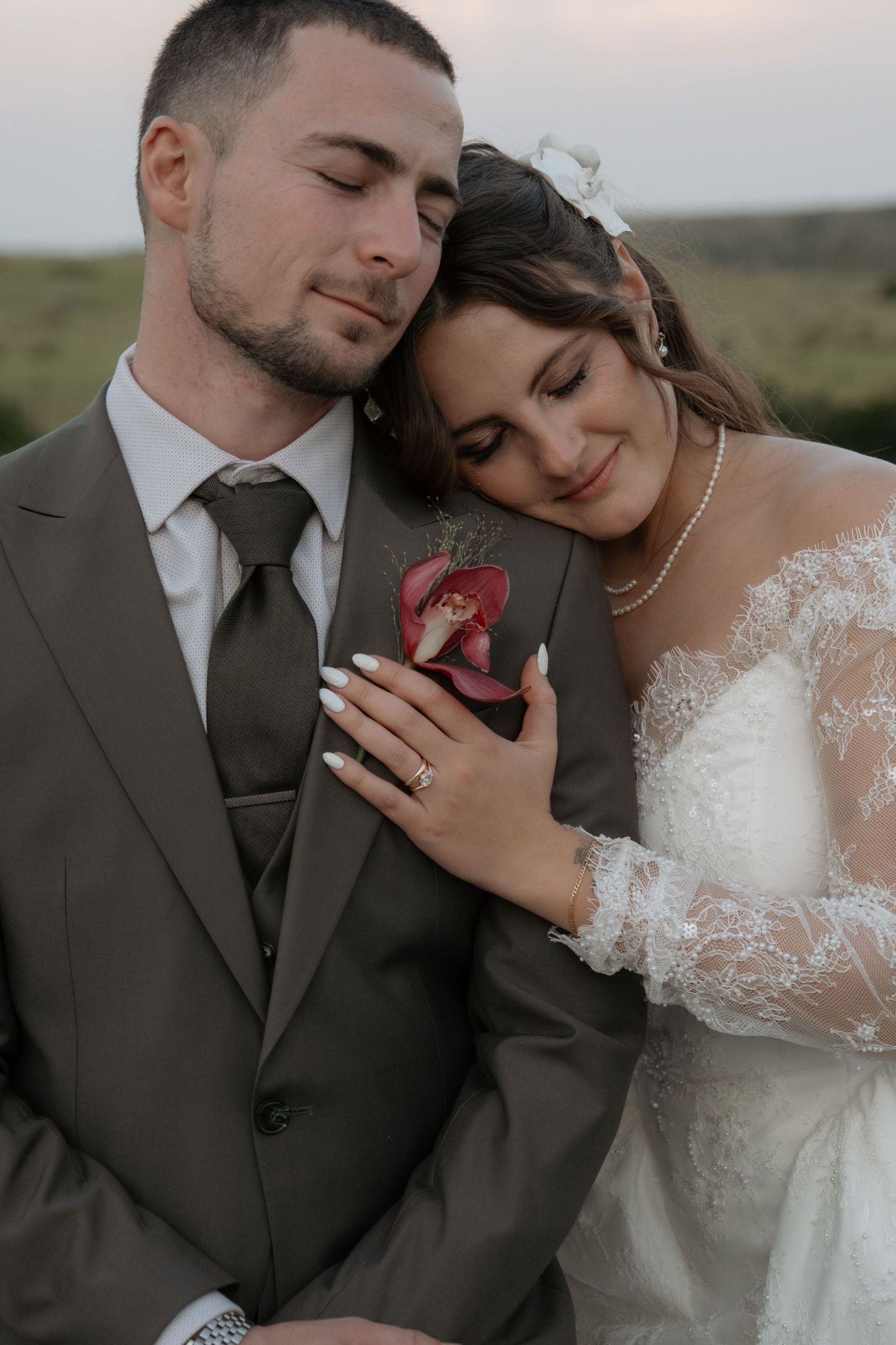 Bride resting head on grooms chest with eyes closed wearing pearl necklace and lace gown.