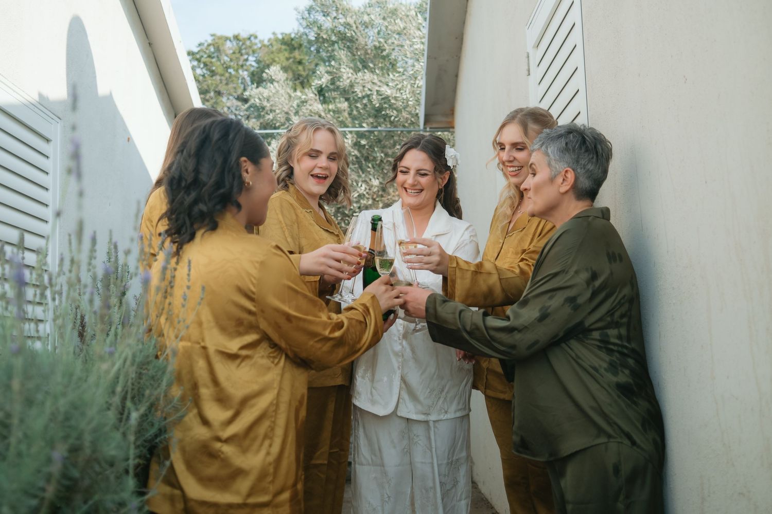 Five people in earthy tones toasting with champagne outdoors, celebrating together near a white wall.