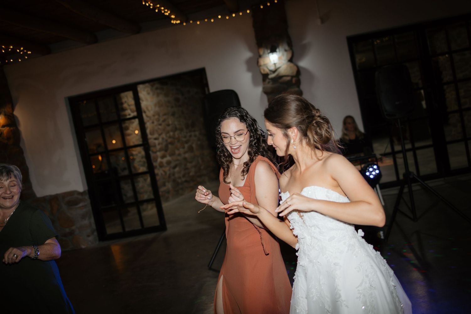 Bride in white strapless gown dances with bridesmaid in rust dress under string lights at wedding reception.