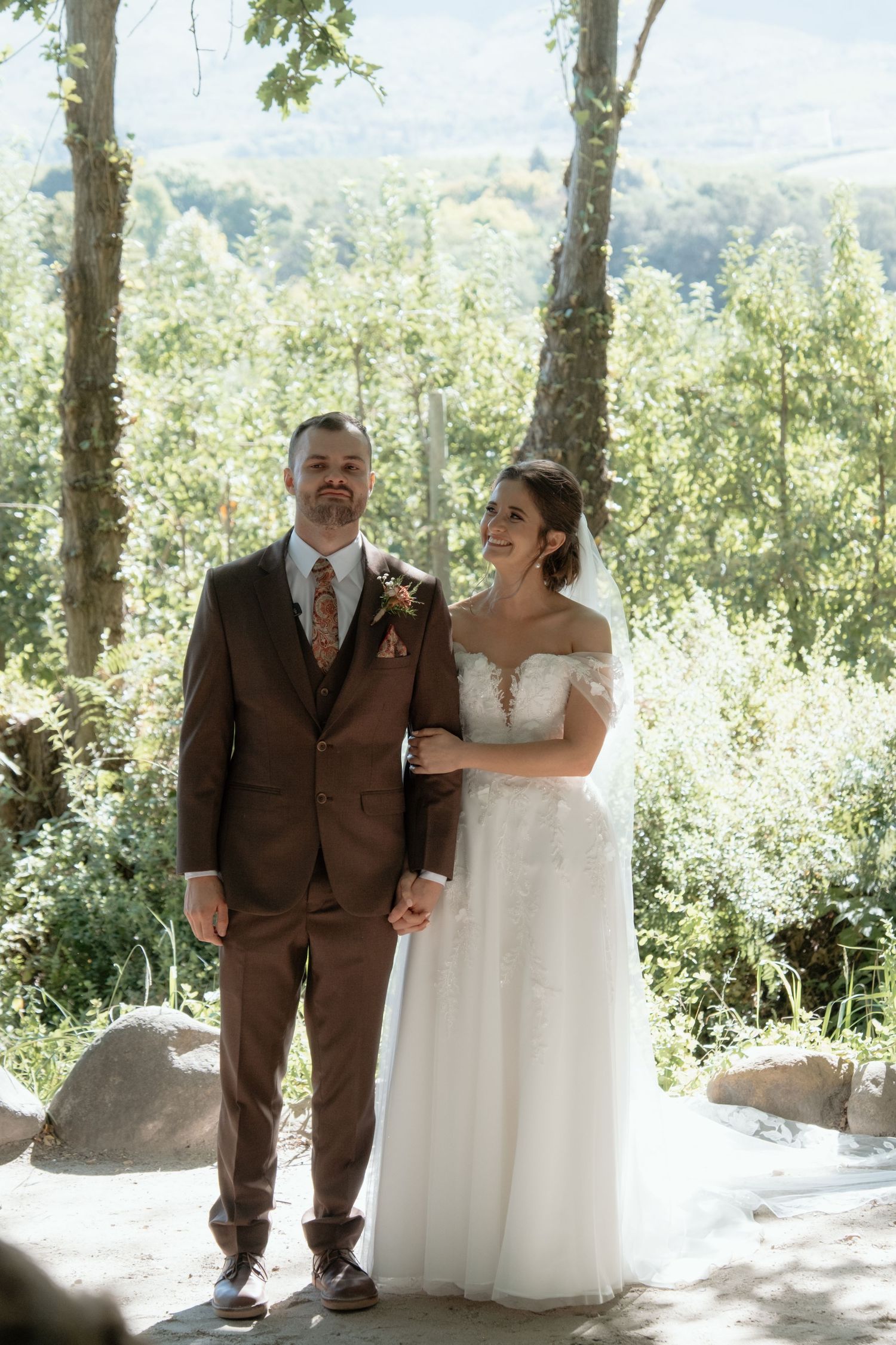 Bride and groom stand together smiling in lush green outdoor wedding setting.