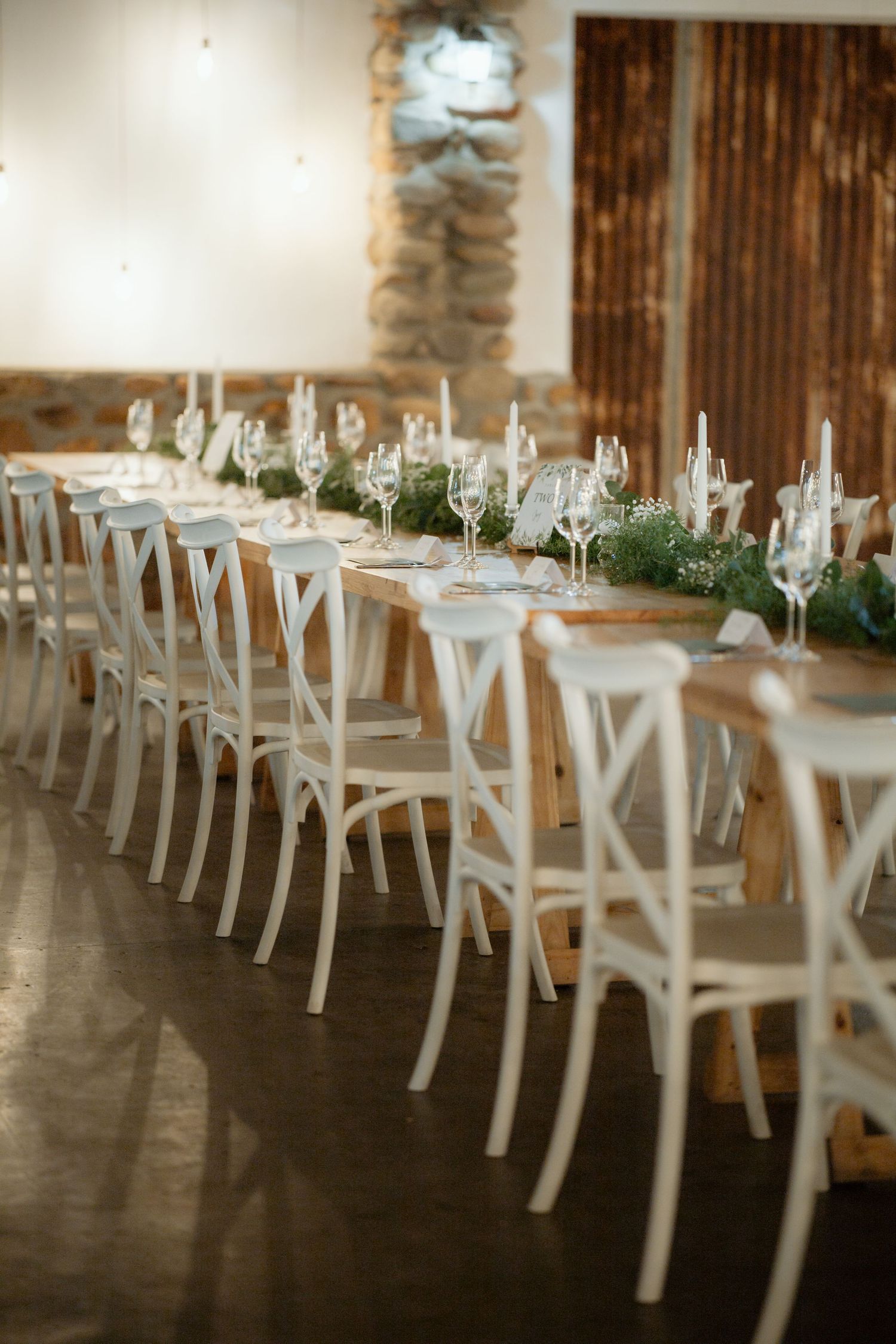 Elegant wedding reception long table with white cross-back chairs, greenery runner, and glassware in a rustic venue.