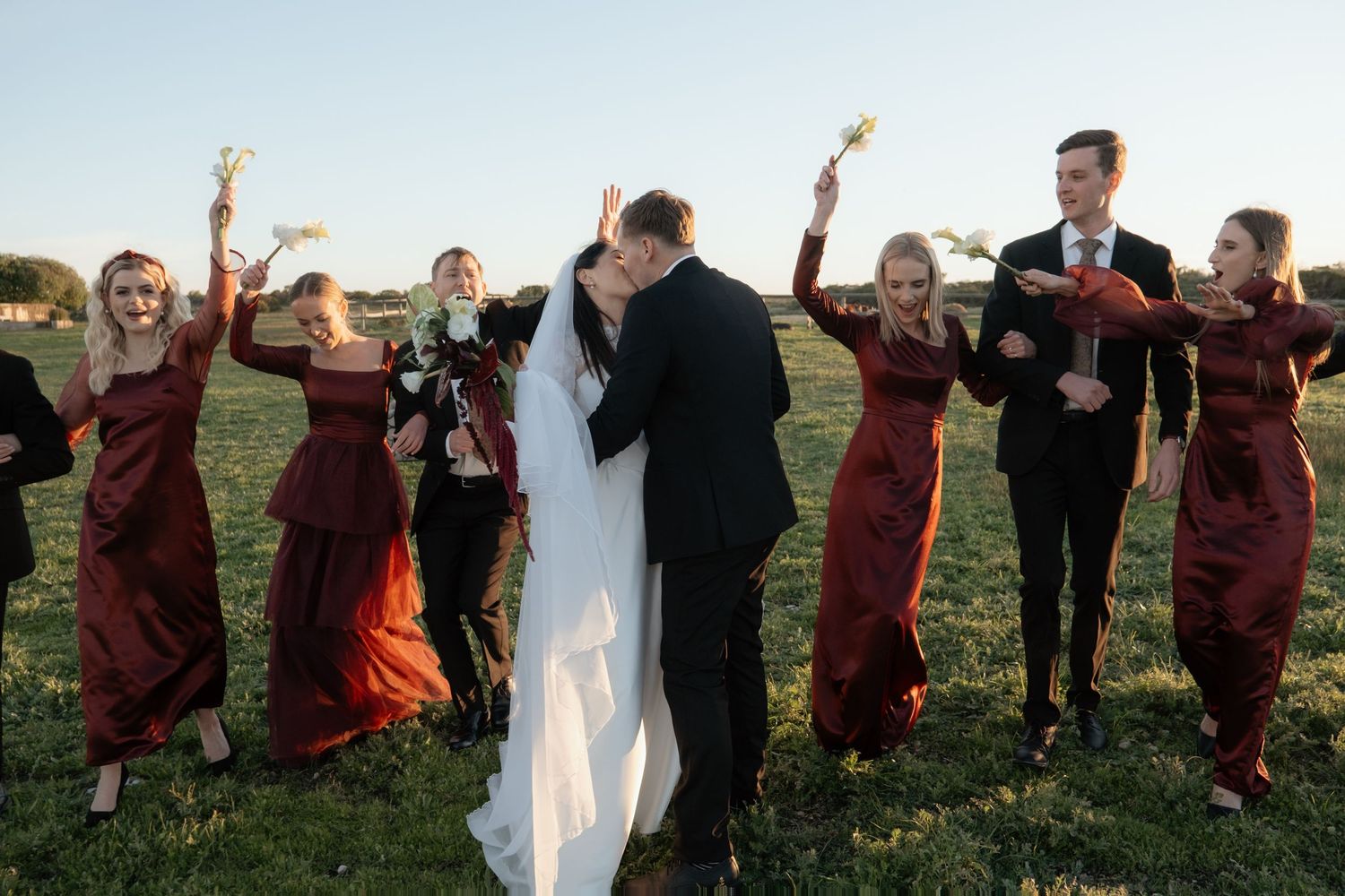 Bride and groom share a romantic kiss surrounded by bridesmaids in burgundy dresses celebrating outdoors.