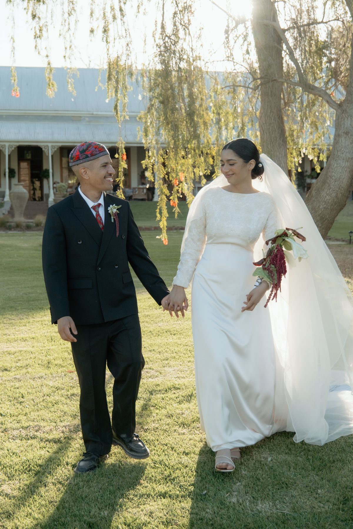 Smiling groom in kufi cap holds brides hand walking across green lawn at outdoor wedding.