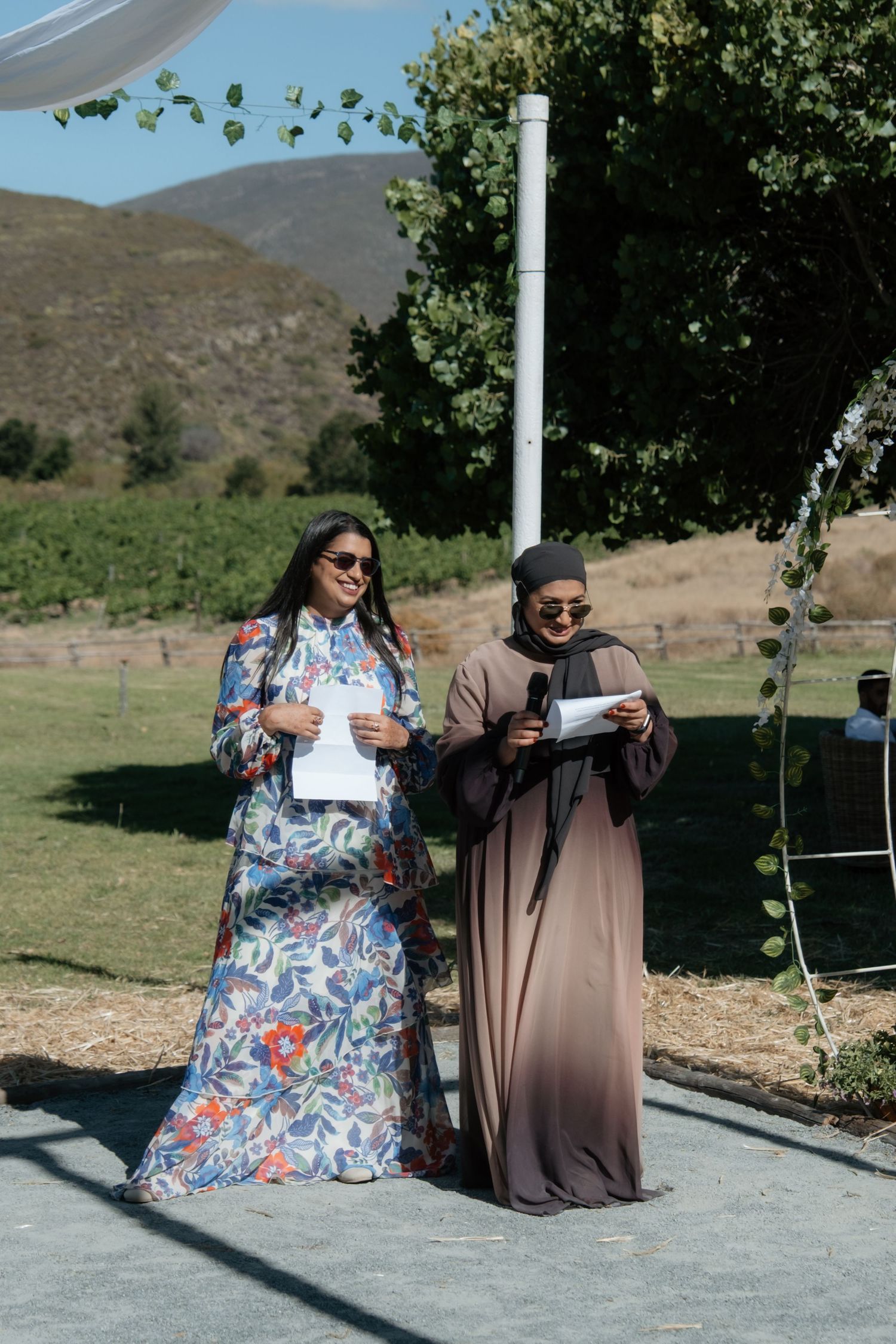 Two women in elegant attire reading speeches at an outdoor wedding ceremony with mountain views.