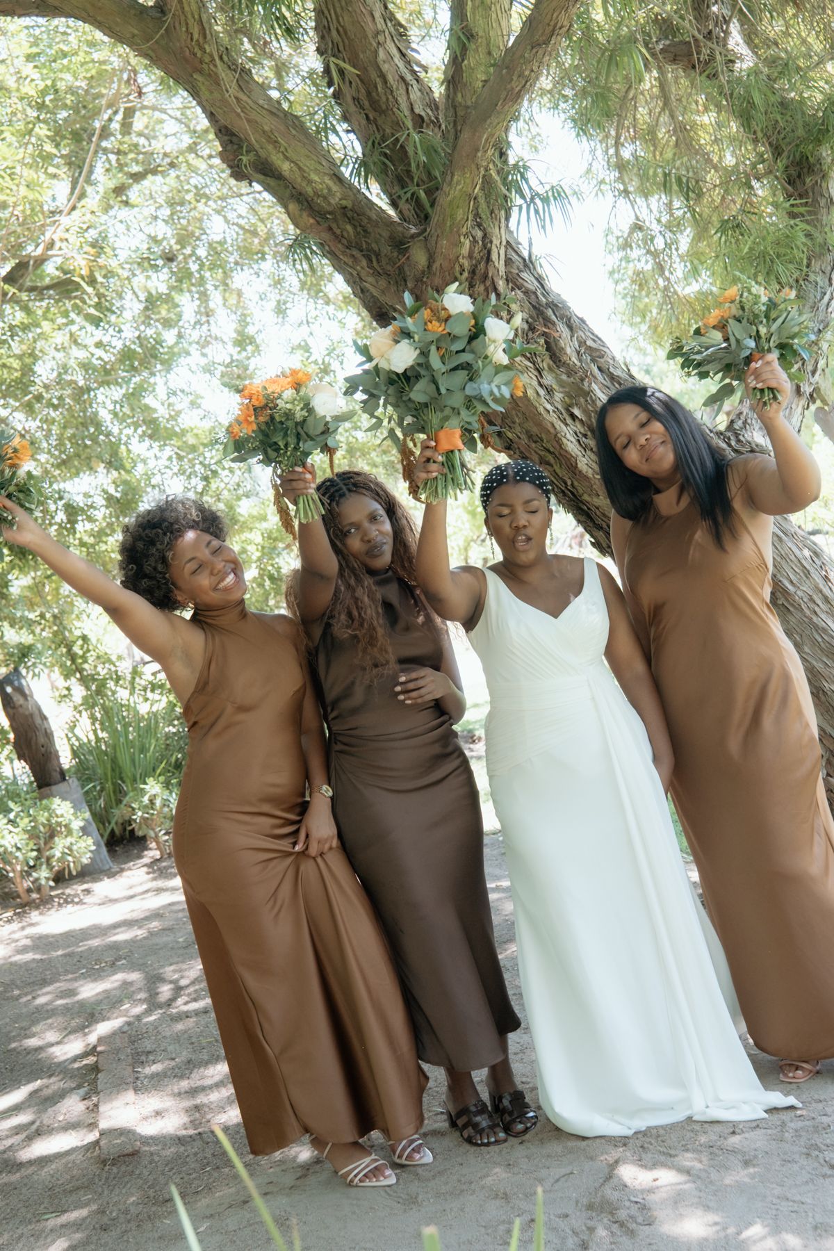Bride in white gown poses with three bridesmaids in brown dresses raising bouquets of orange and white flowers.