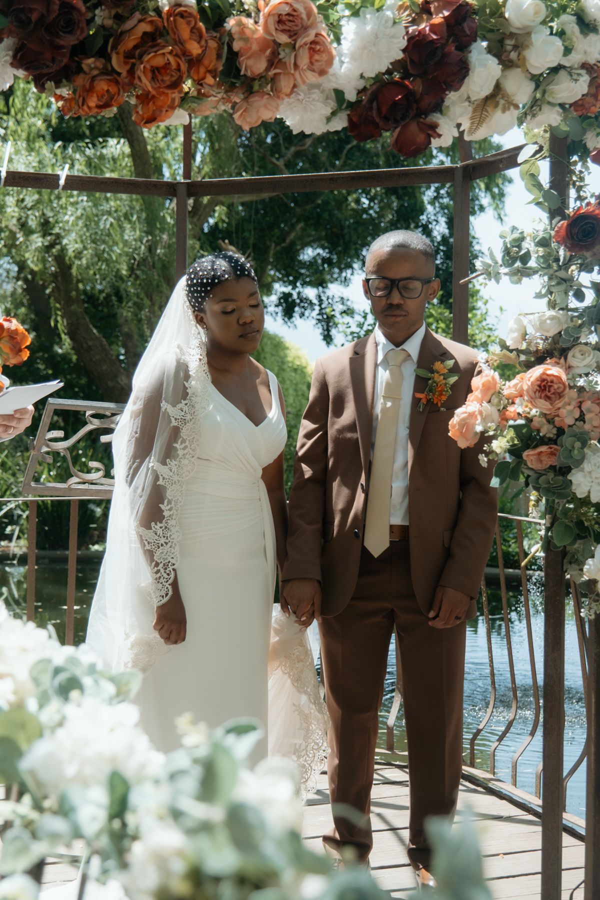 Bride in white lace veil and groom in brown suit hold hands under floral arch.