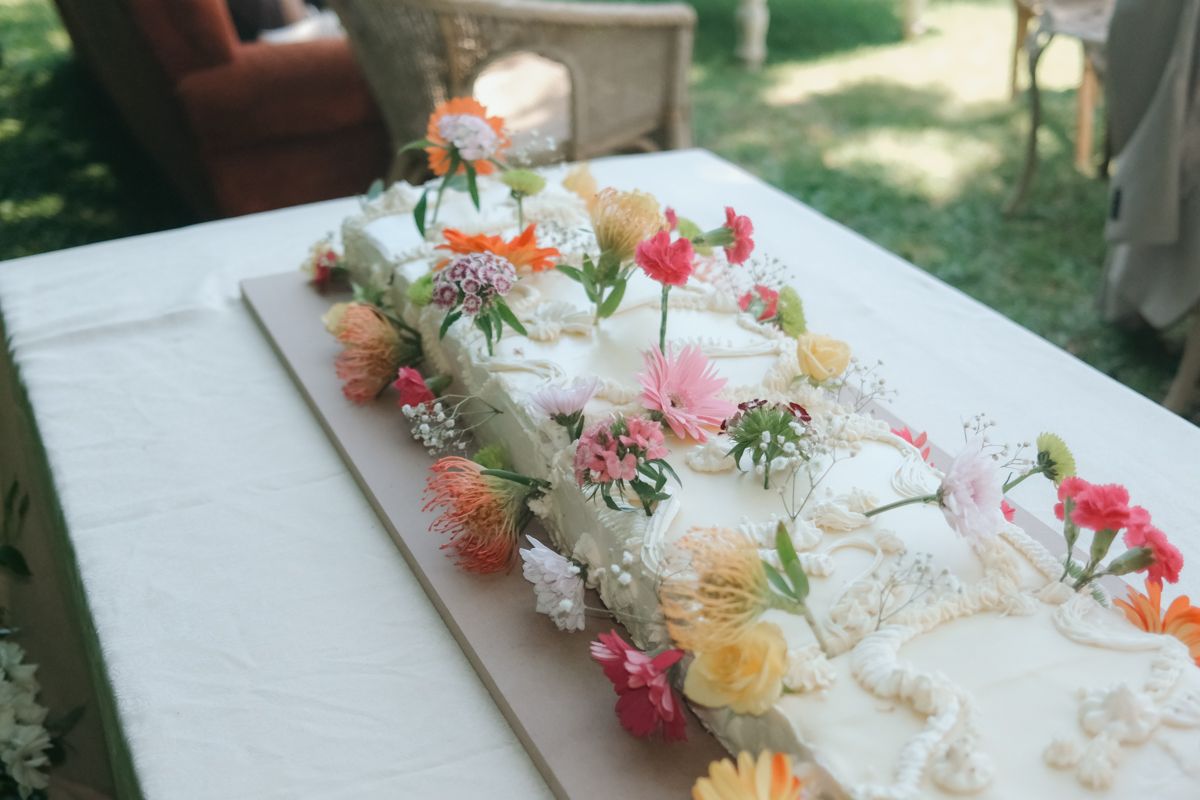 Elegant wedding cake decorated with colorful fresh flowers including pink, orange, and yellow blooms on a white tablecloth.