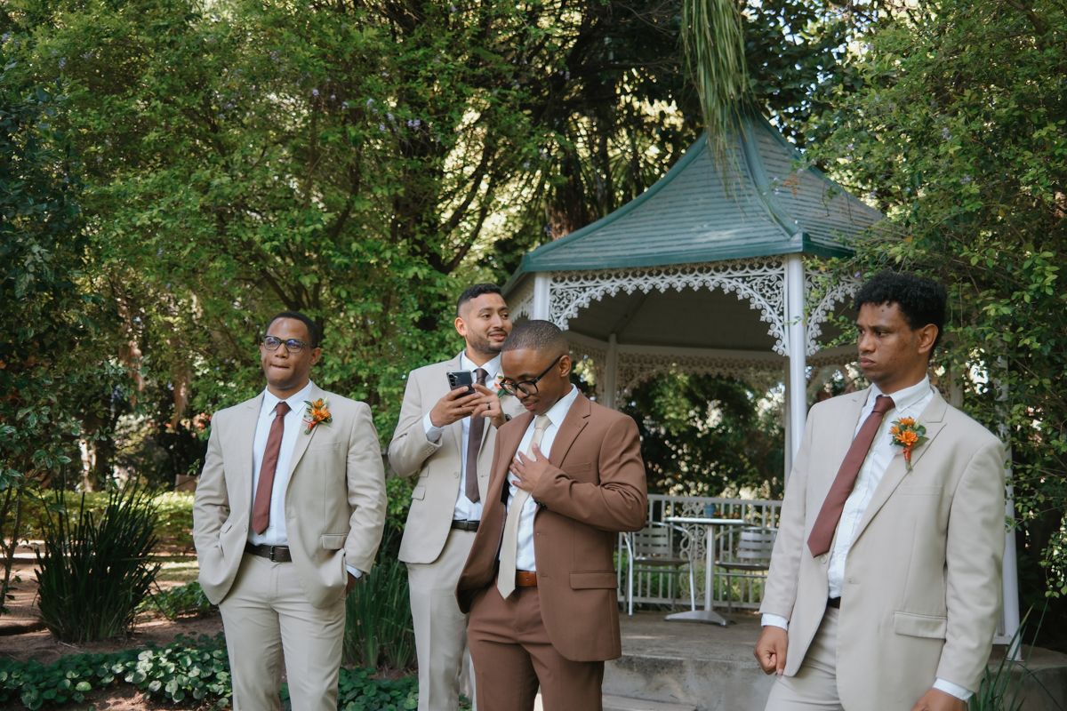 Four groomsmen in beige and brown wedding suits stand casually near a decorative white gazebo outdoors.
