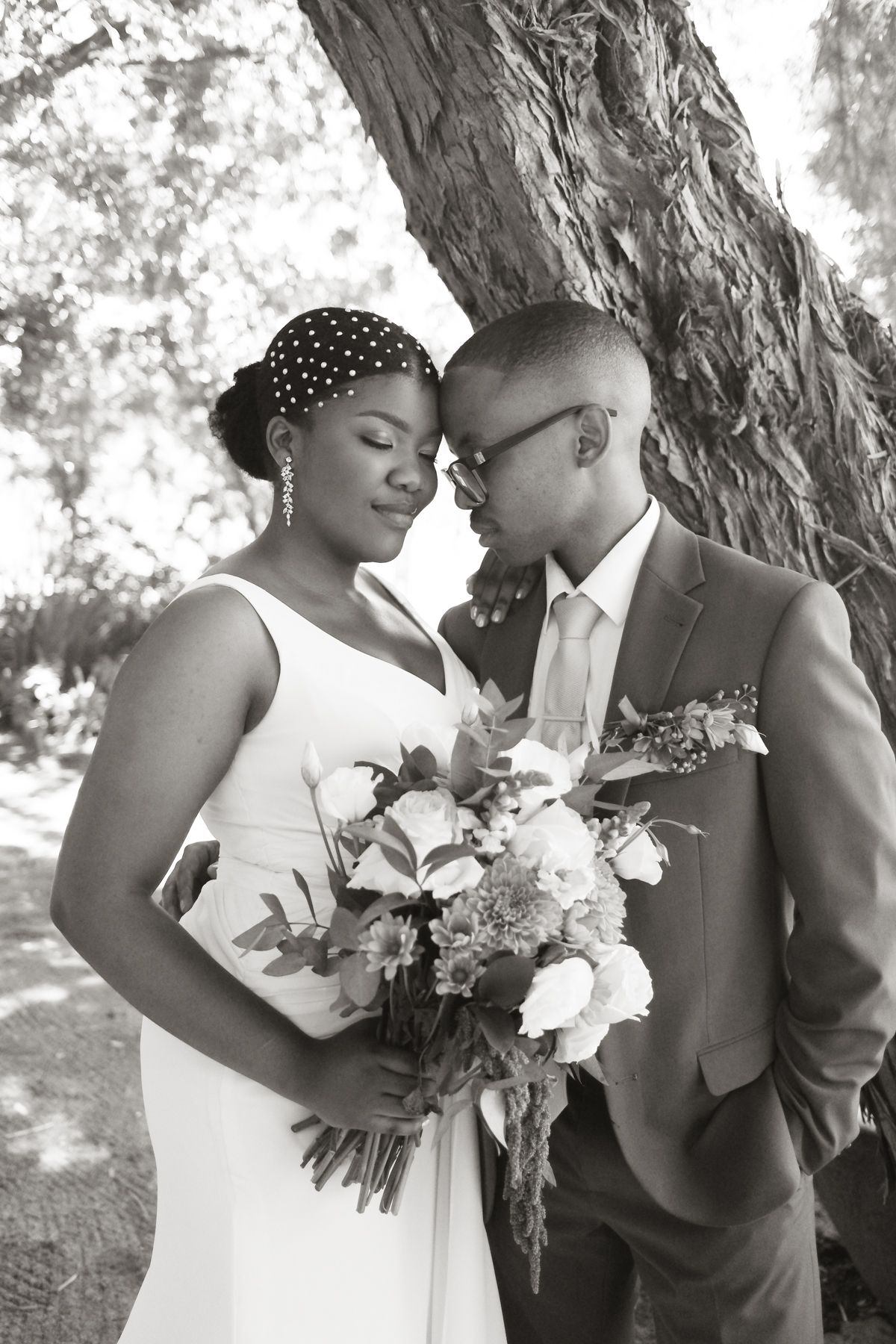 Black and white portrait of an elegant bride and groom embracing near a tree, bride holding a bouquet.