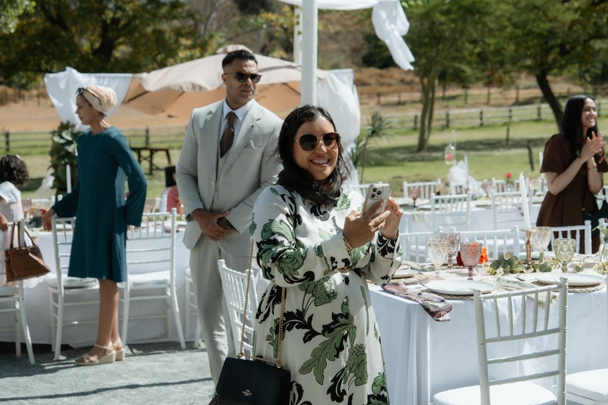 Wedding guest in floral dress taking a selfie at an elegant outdoor reception with white table settings.