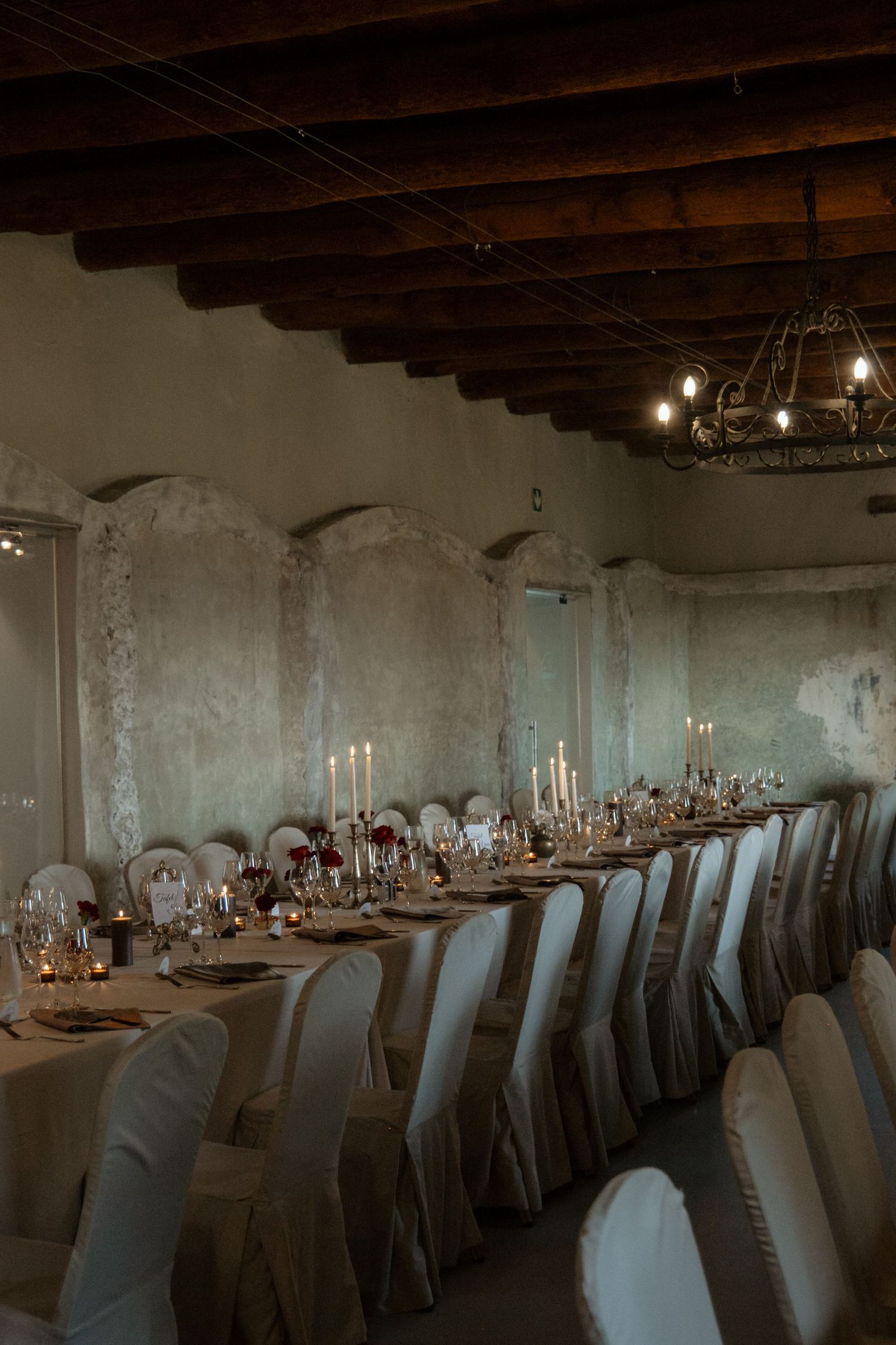Elegant wedding reception hall with long candlelit table, white chair covers, and rustic wooden ceiling in historic venue.