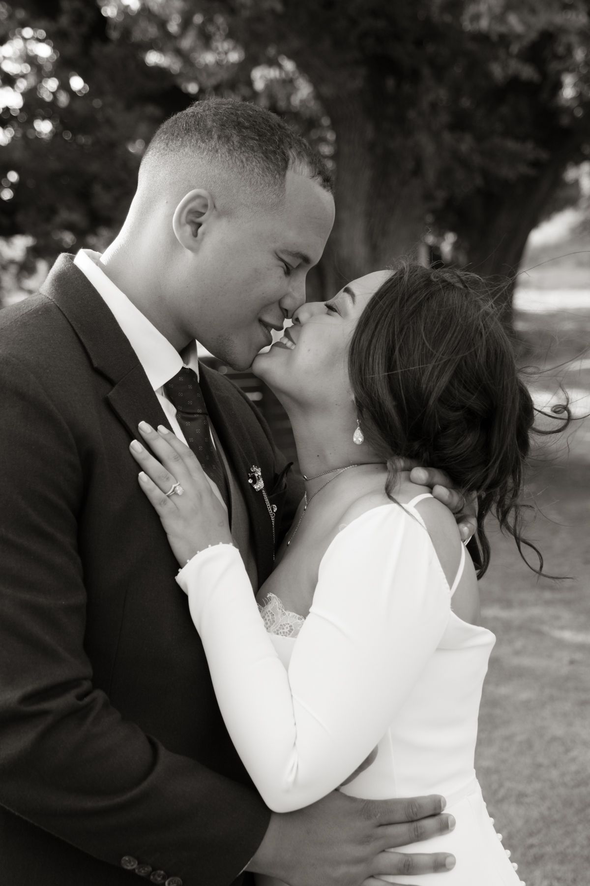 Black and white romantic portrait of bride and groom sharing a kiss outdoors under trees.