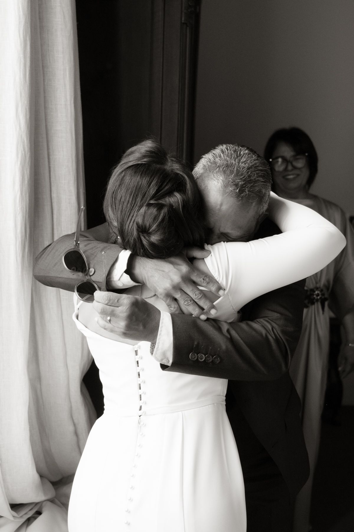 Black and white photo of a bride in a white gown sharing an emotional hug with a family member before the ceremony.