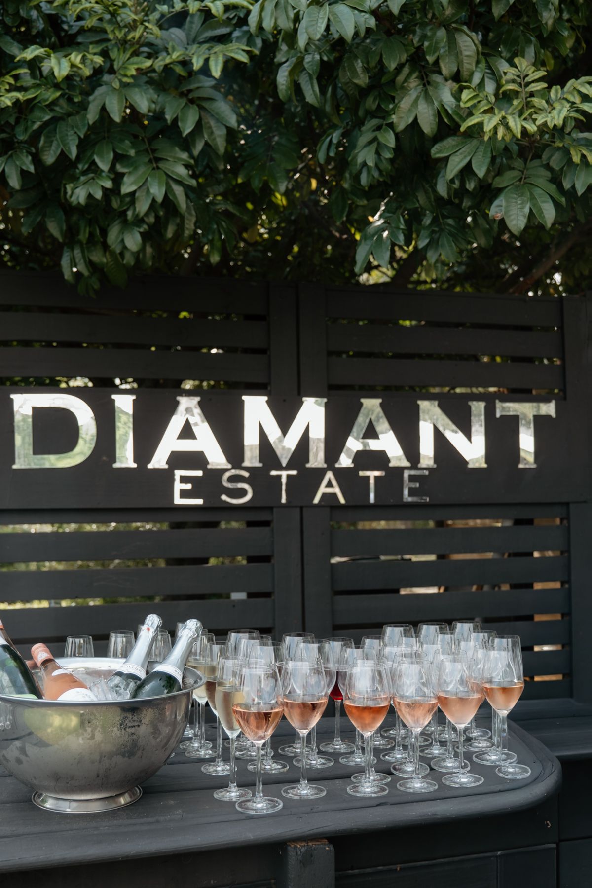 Diamant Estate wedding venue sign with rose champagne glasses and bottle display on outdoor table.