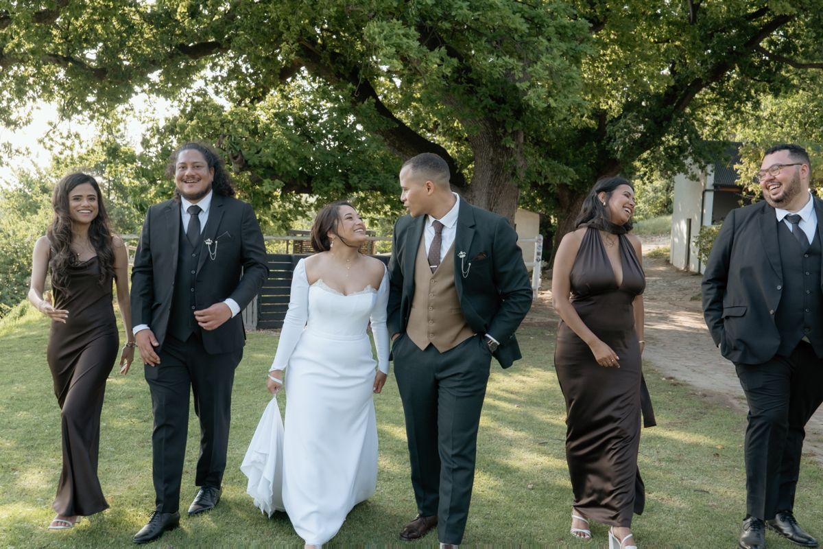 Bride and groom walk with wedding party in brown attire under large tree at outdoor venue.