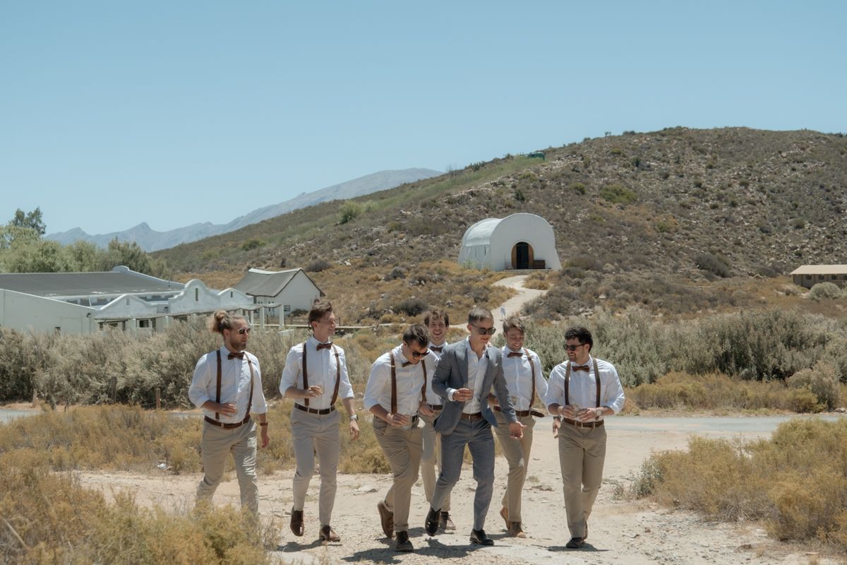 Groom with groomsmen wearing suspenders and khaki pants posing in arid landscape with dome building on hillside.