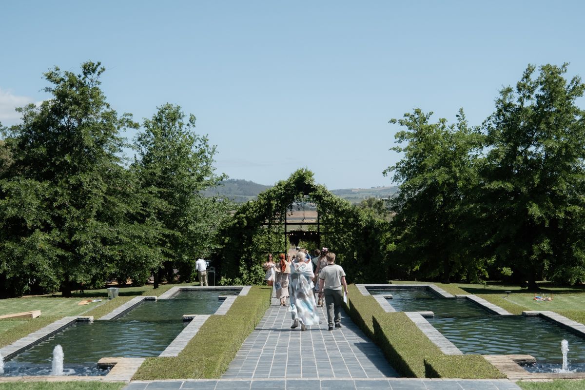 Wedding party walking along elegant garden path with reflecting pools toward ivy-covered pergola backdrop.