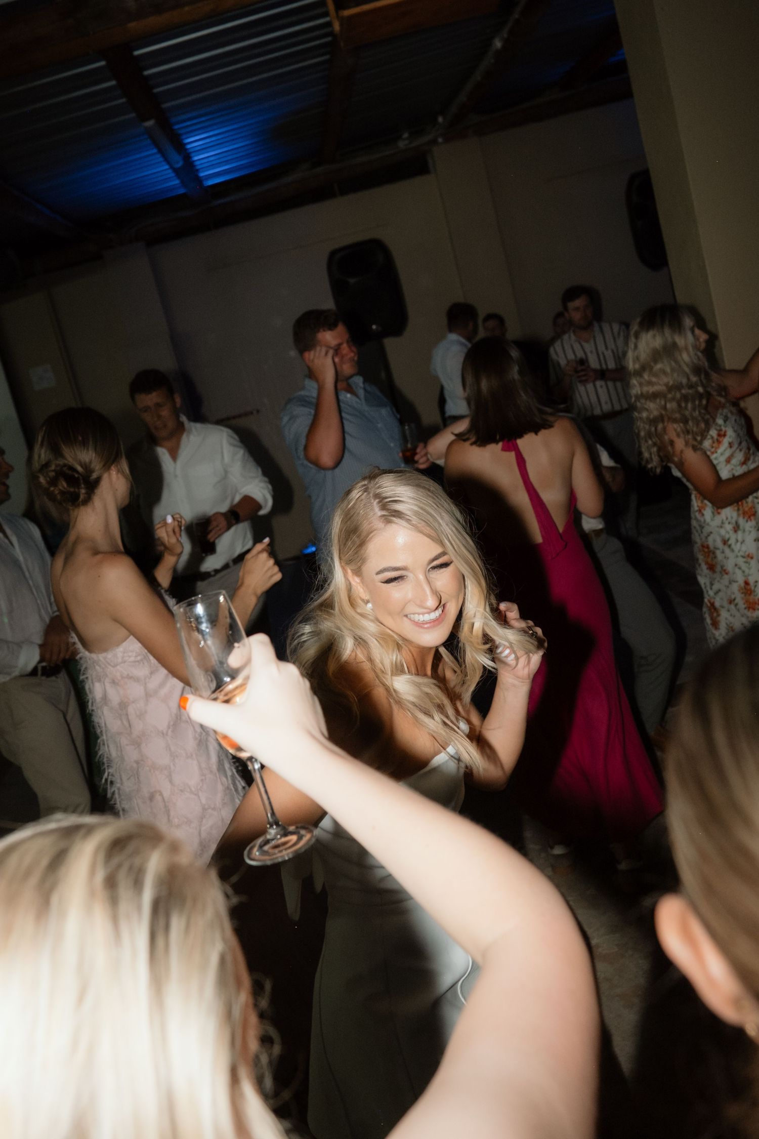Blonde wedding guest smiling and dancing with champagne glass at lively indoor wedding reception dance floor.