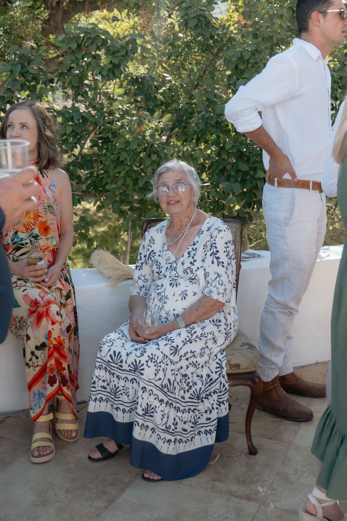 Elderly woman in floral dress seated smiling at outdoor wedding reception among guests.