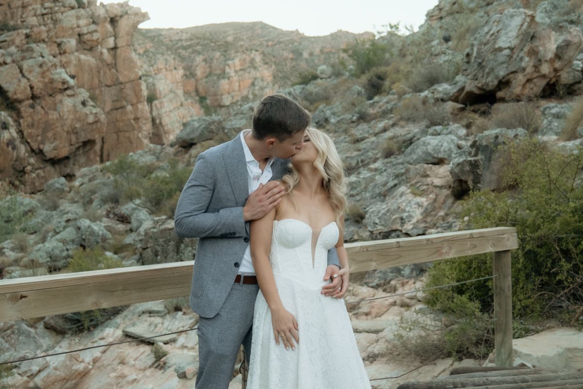 Bride and groom sharing a kiss on a wooden bridge with rocky mountain landscape in background.