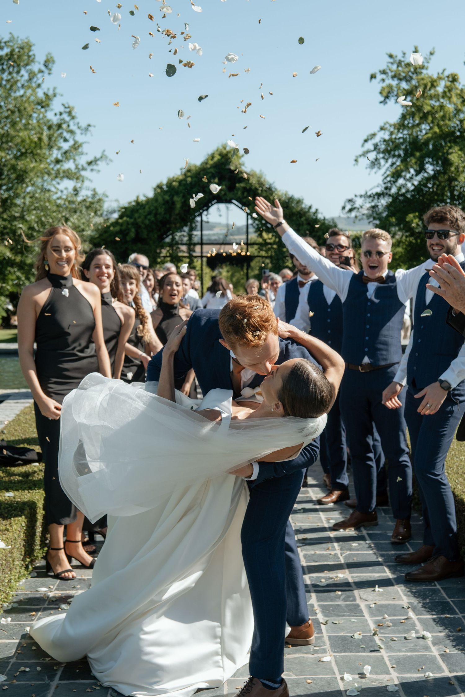Groom dips bride for a kiss while guests throw confetti during joyful outdoor wedding ceremony exit.