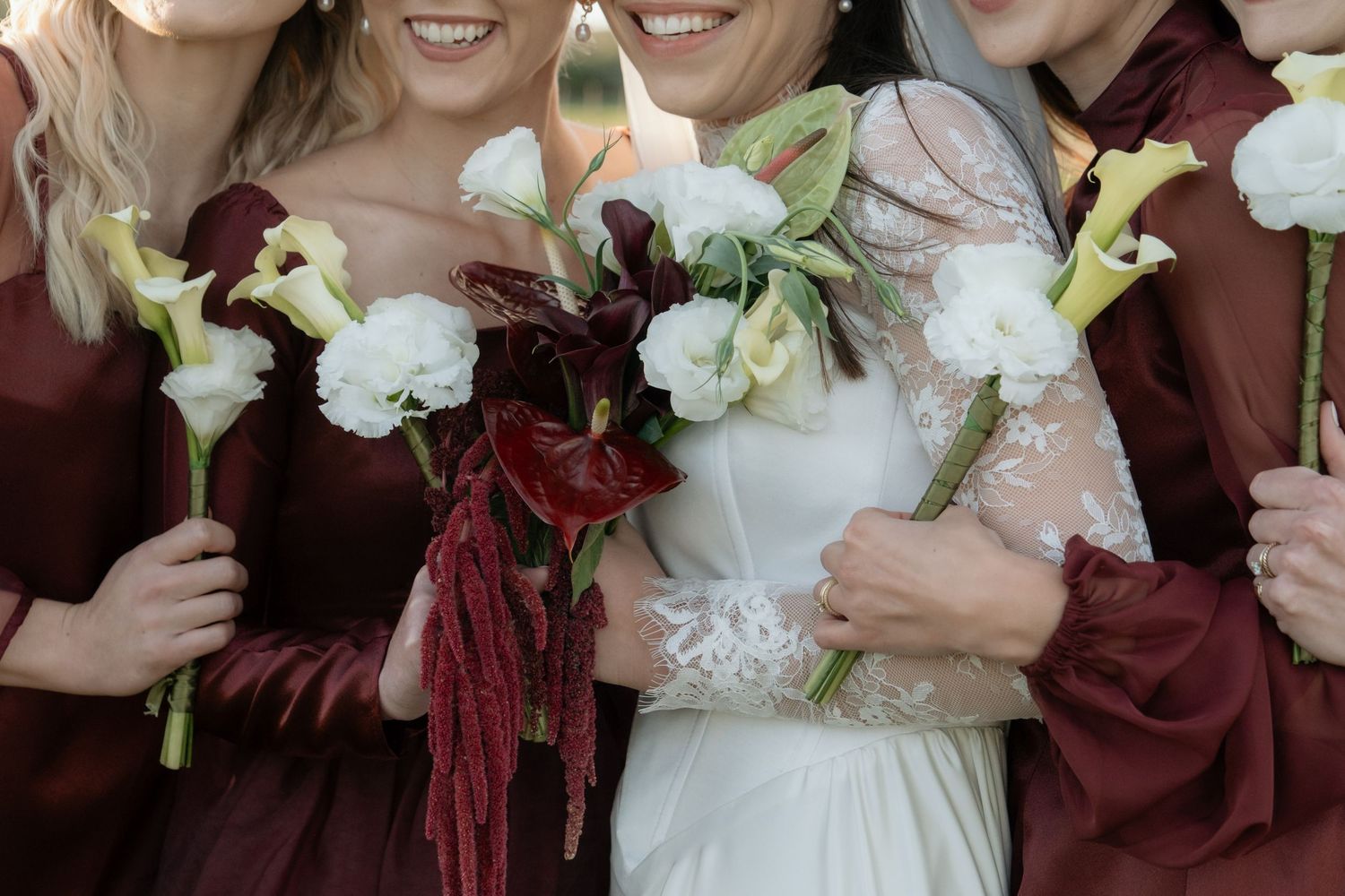 Bride and bridesmaids in burgundy dresses holding white calla lily and rose bouquets at a wedding ceremony.