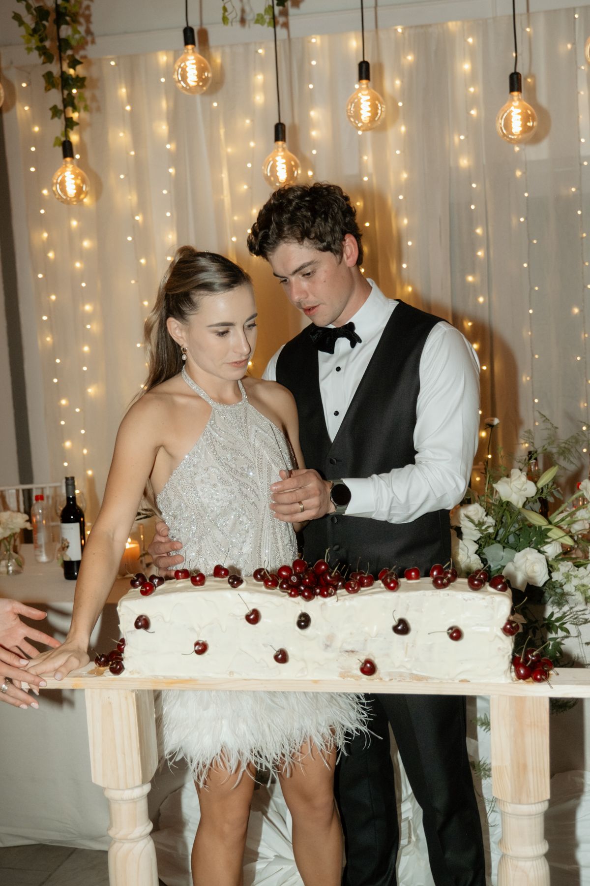 Bride and groom in formal attire cutting their white wedding cake decorated with cherries at reception.