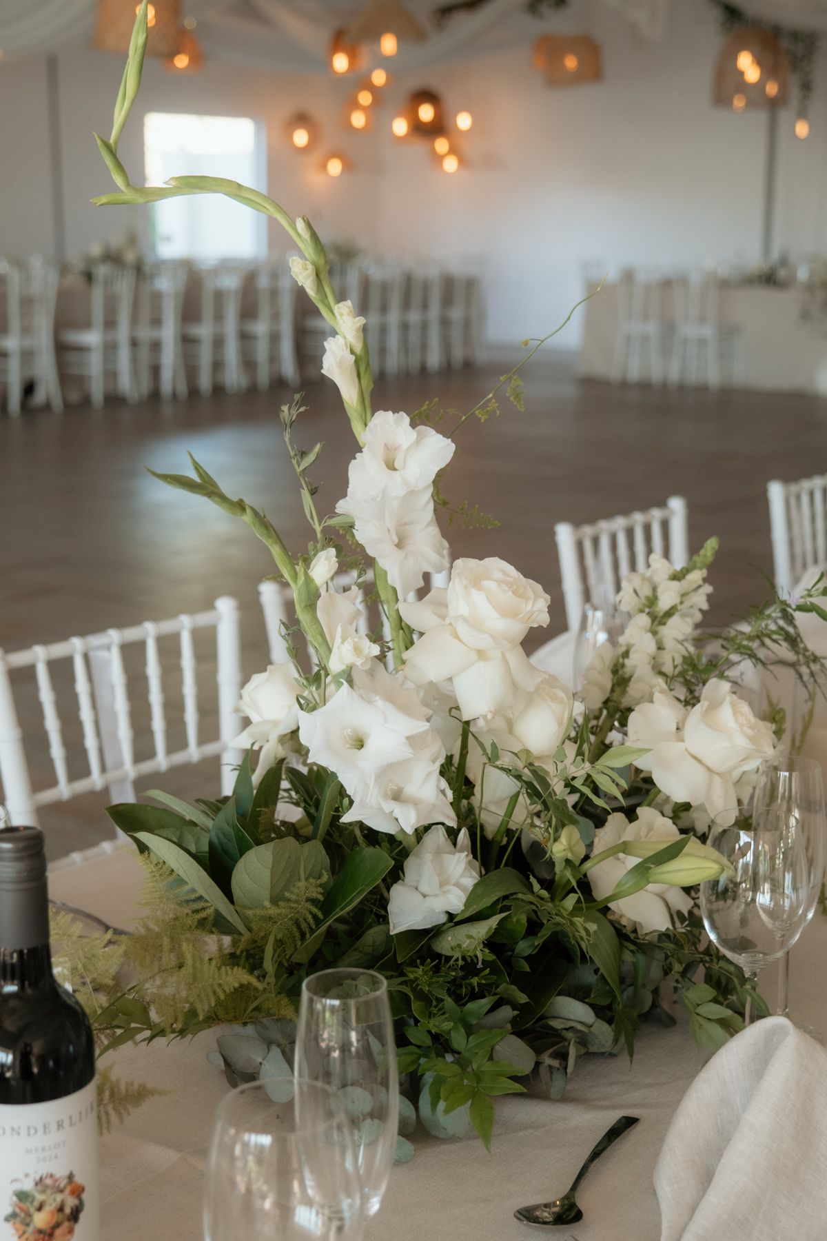 Elegant wedding reception centerpiece with white roses, gladiolus, and greenery on a decorated table in a bright venue.