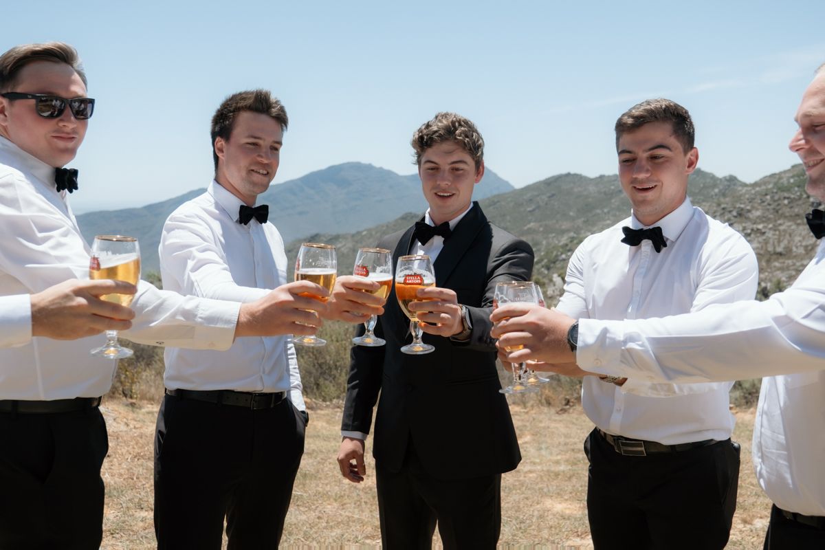 Groomsmen in white shirts and bow ties toasting with drinks outdoors against a scenic mountain backdrop.