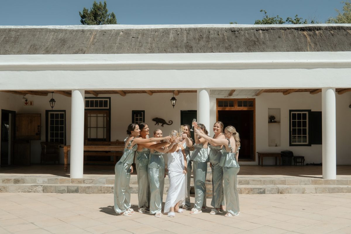 A bride and bridesmaids in sage green dresses celebrating together outside a white colonial-style venue.
