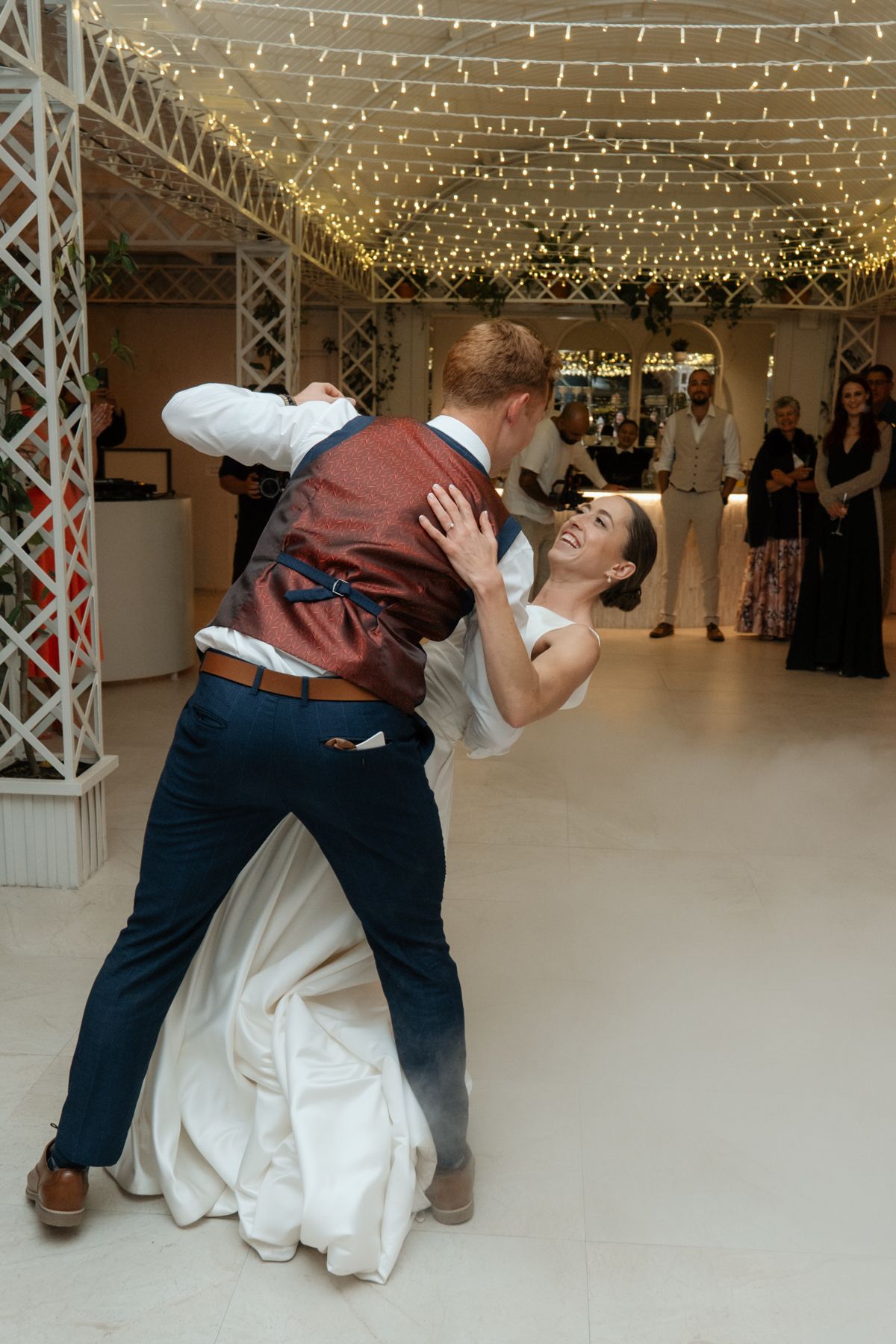Bride laughing joyfully as groom dips her during their first dance under twinkling fairy lights on the dance floor.