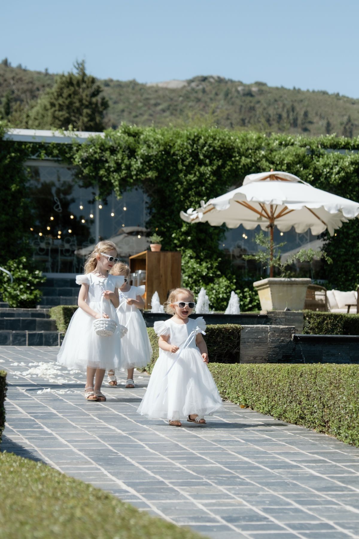 Three young flower girls in white dresses and sunglasses walking along a garden pathway.