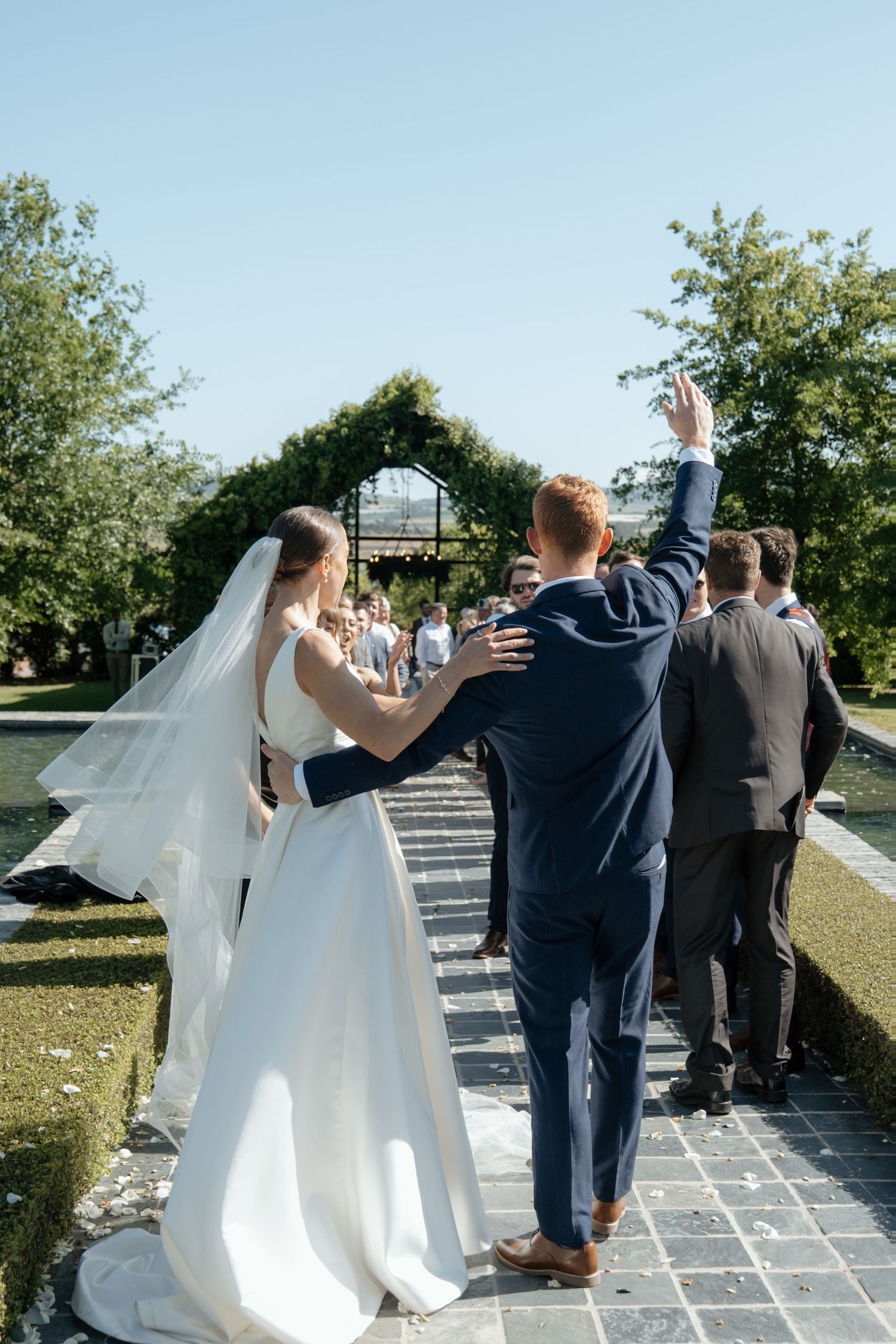 Newlywed couple walks down aisle as groom raises hand to celebrate after wedding ceremony.