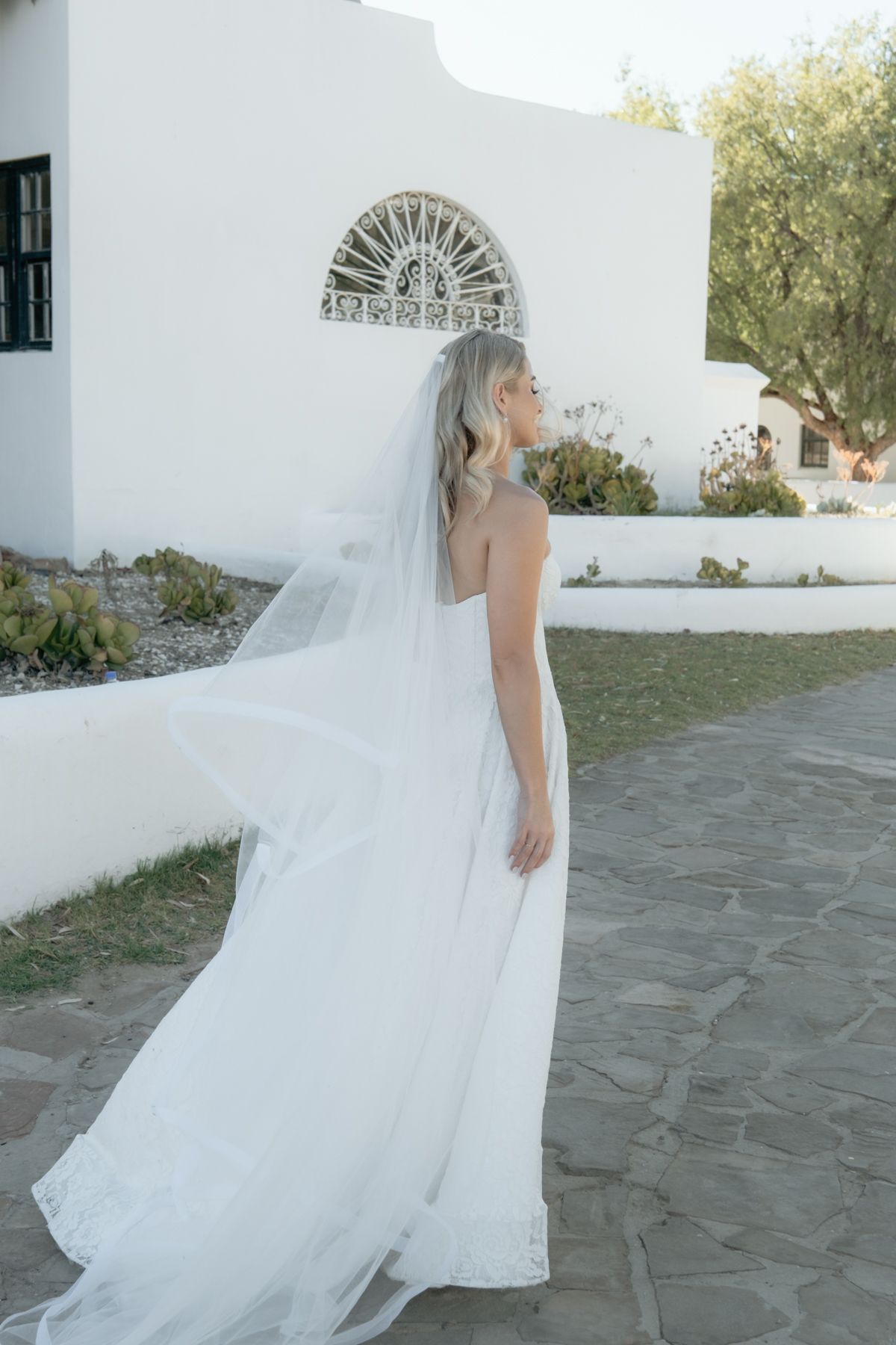 Bride in a flowing white wedding gown and veil stands on stone path before a white chapel.