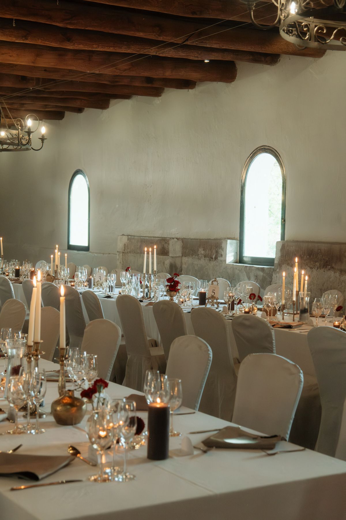 Elegant wedding reception hall with long tables, white linens, tall candles, and rustic wooden beam ceiling.