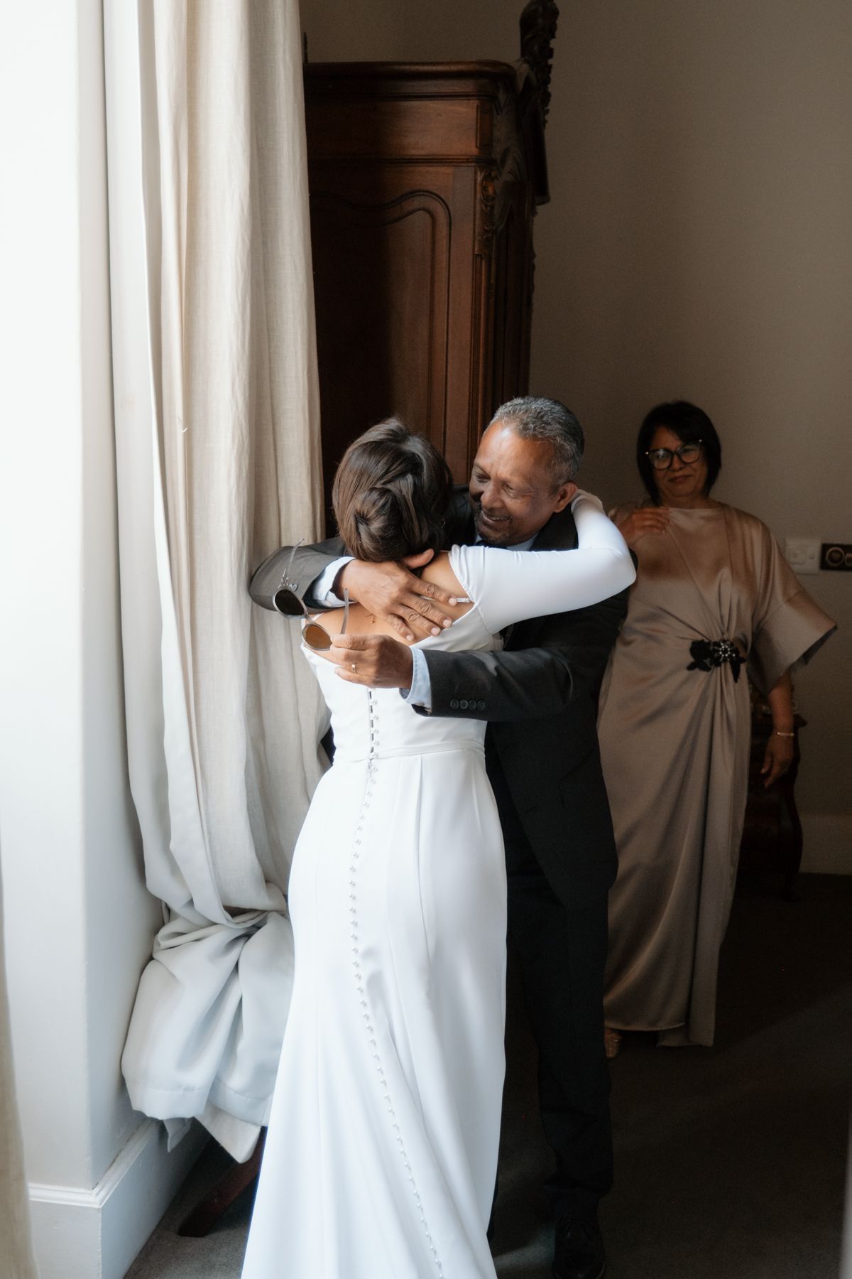 Bride embracing an older gentleman warmly while a woman watches in the background.