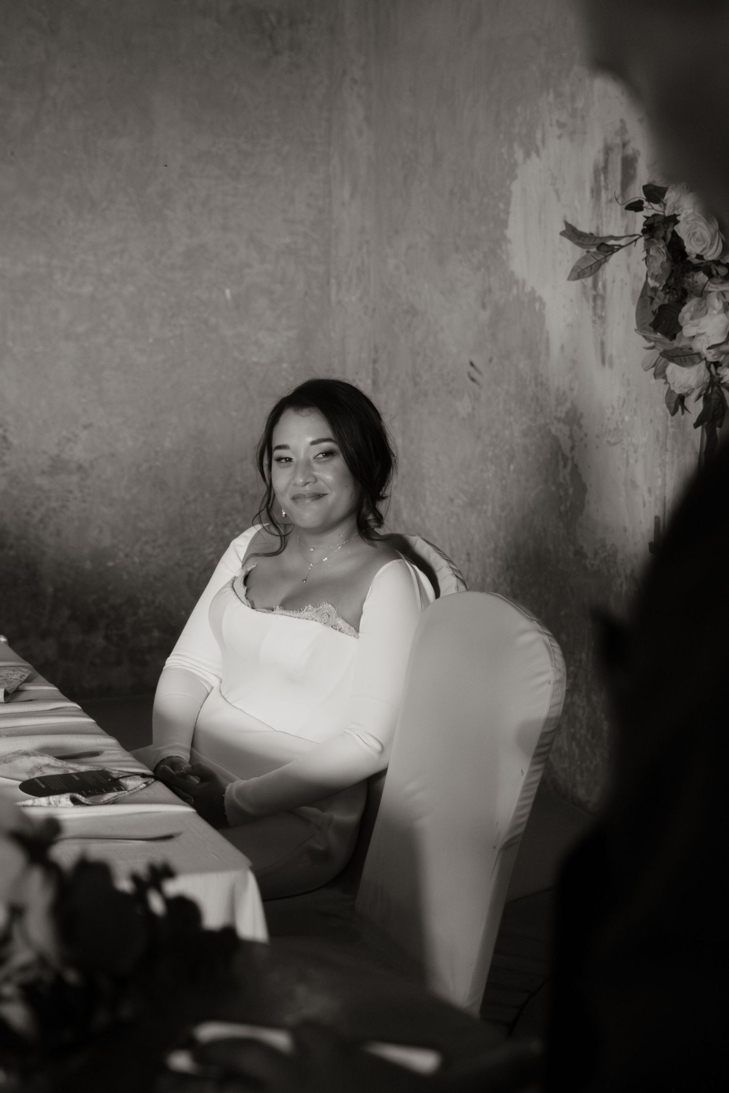 Black and white portrait of a smiling bride in white dress seated at wedding reception dinner table.