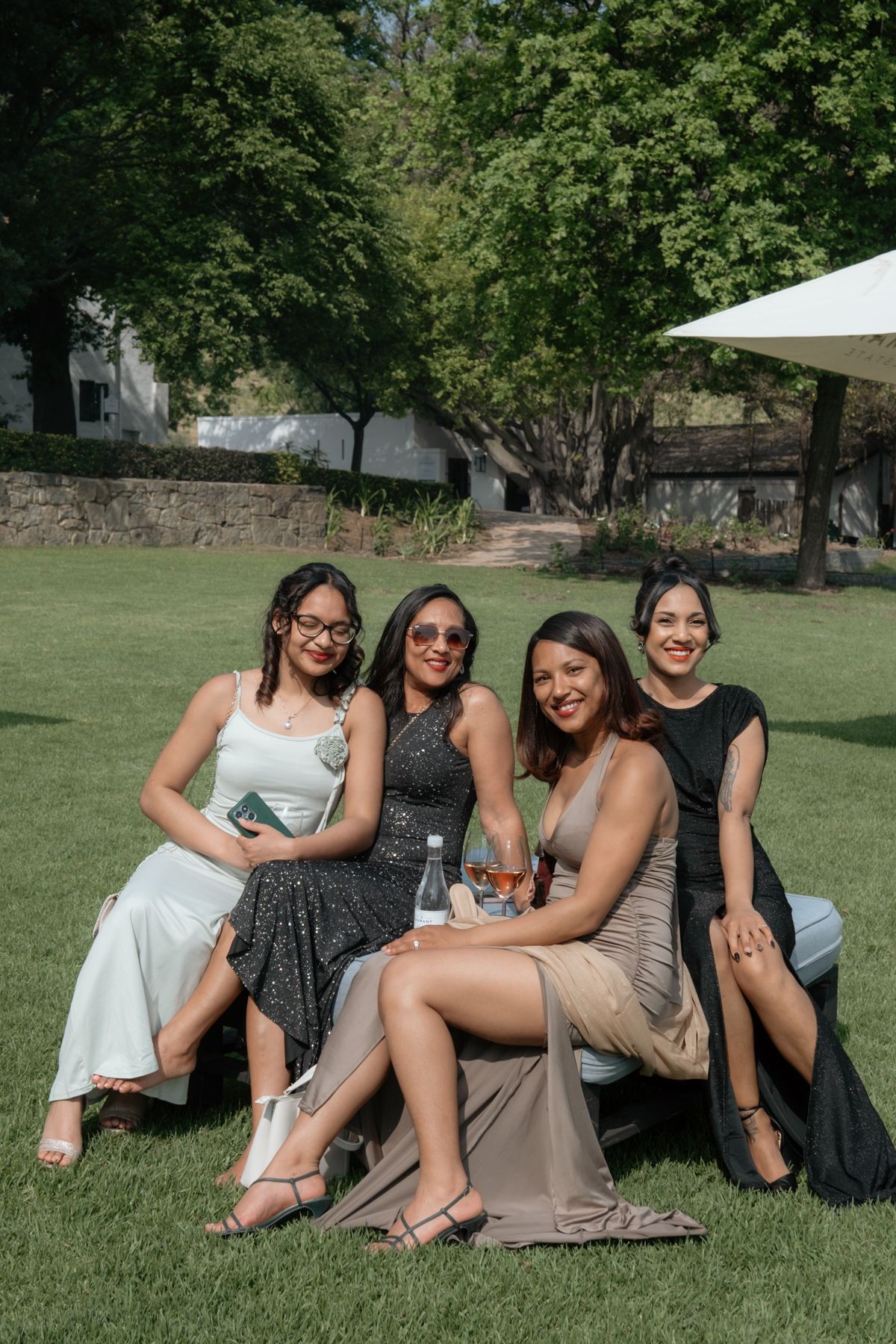 Four elegantly dressed women posing together on outdoor seating at a sunny garden wedding celebration.